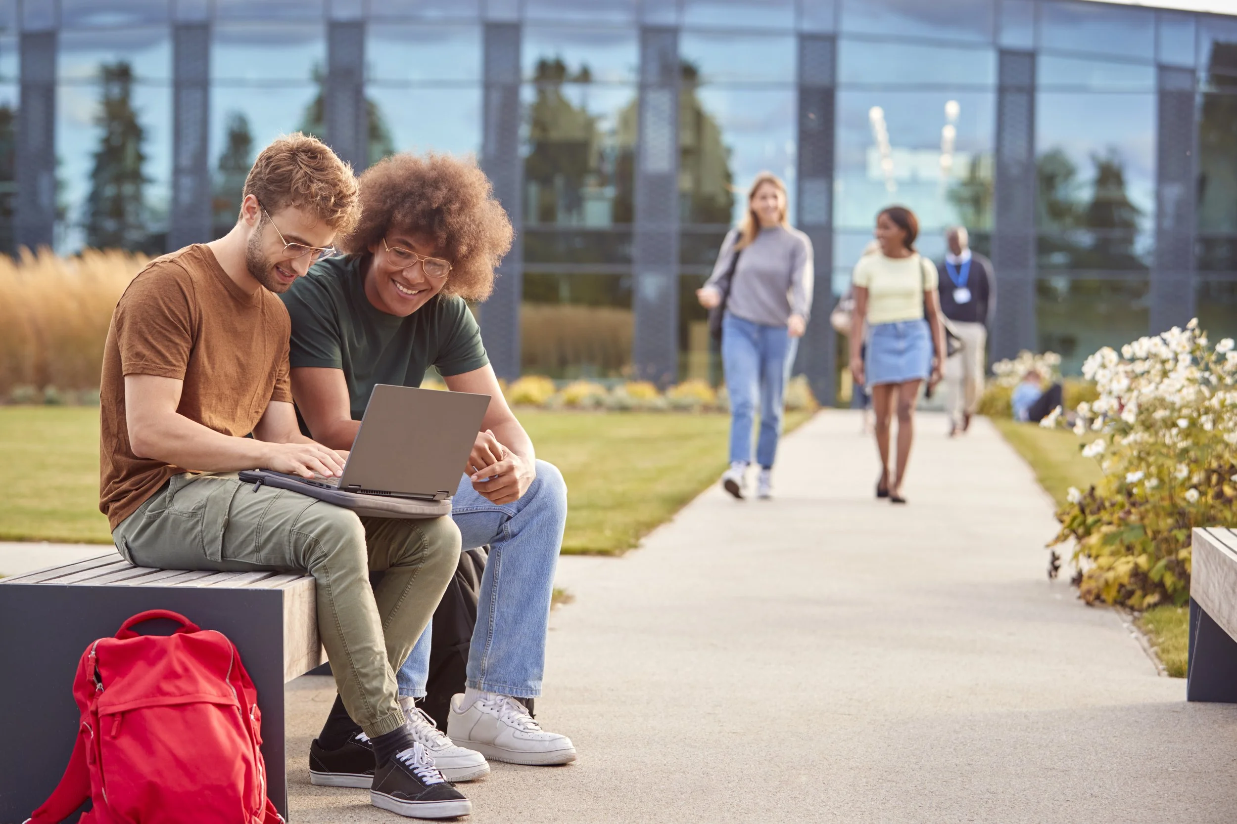Two college students sitting on a bench outdoors, looking at a laptop and smiling, with a red backpack nearby, while others walk on a sidewalk in front of a modern glass building.