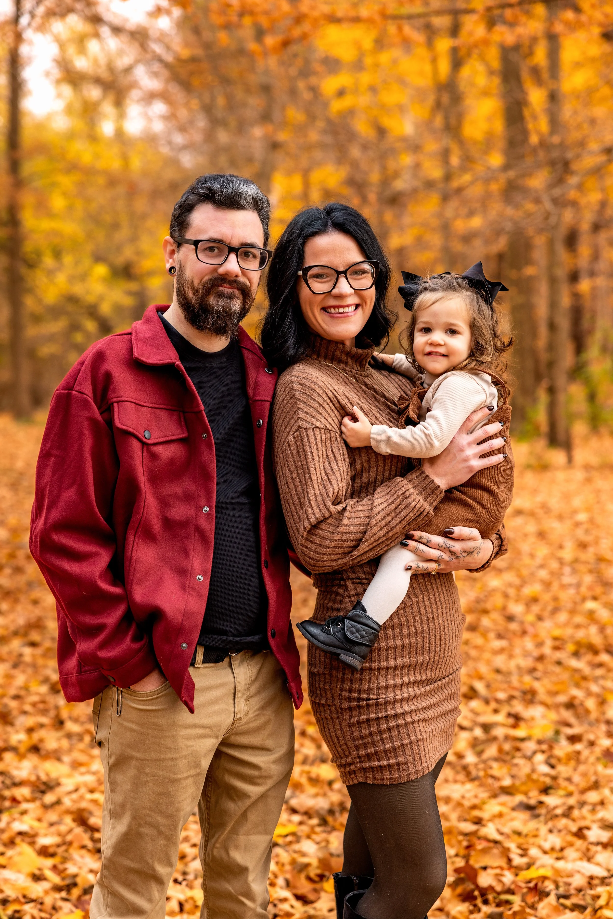A smiling family of three standing outdoors in an autumn forest with colorful fall leaves on the trees and ground, posing for a photo.
