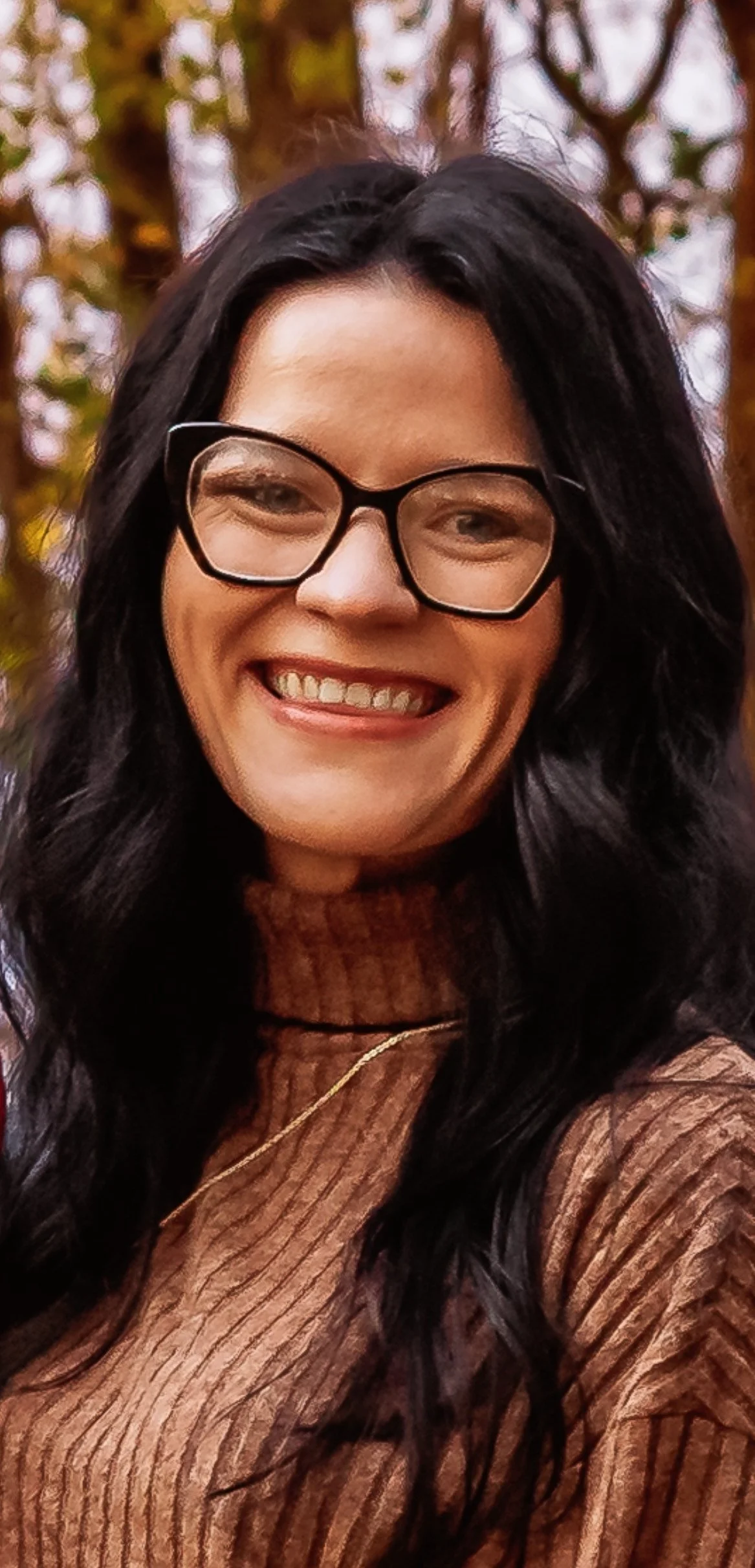 A woman with black wavy hair wearing glasses and a brown ribbed turtleneck sweater, smiling outdoors with blurred autumn trees in the background.