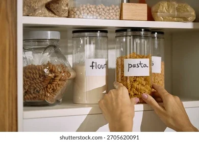 Person placing pasta into labeled glass jar on kitchen shelf with other jars containing flour and rice.