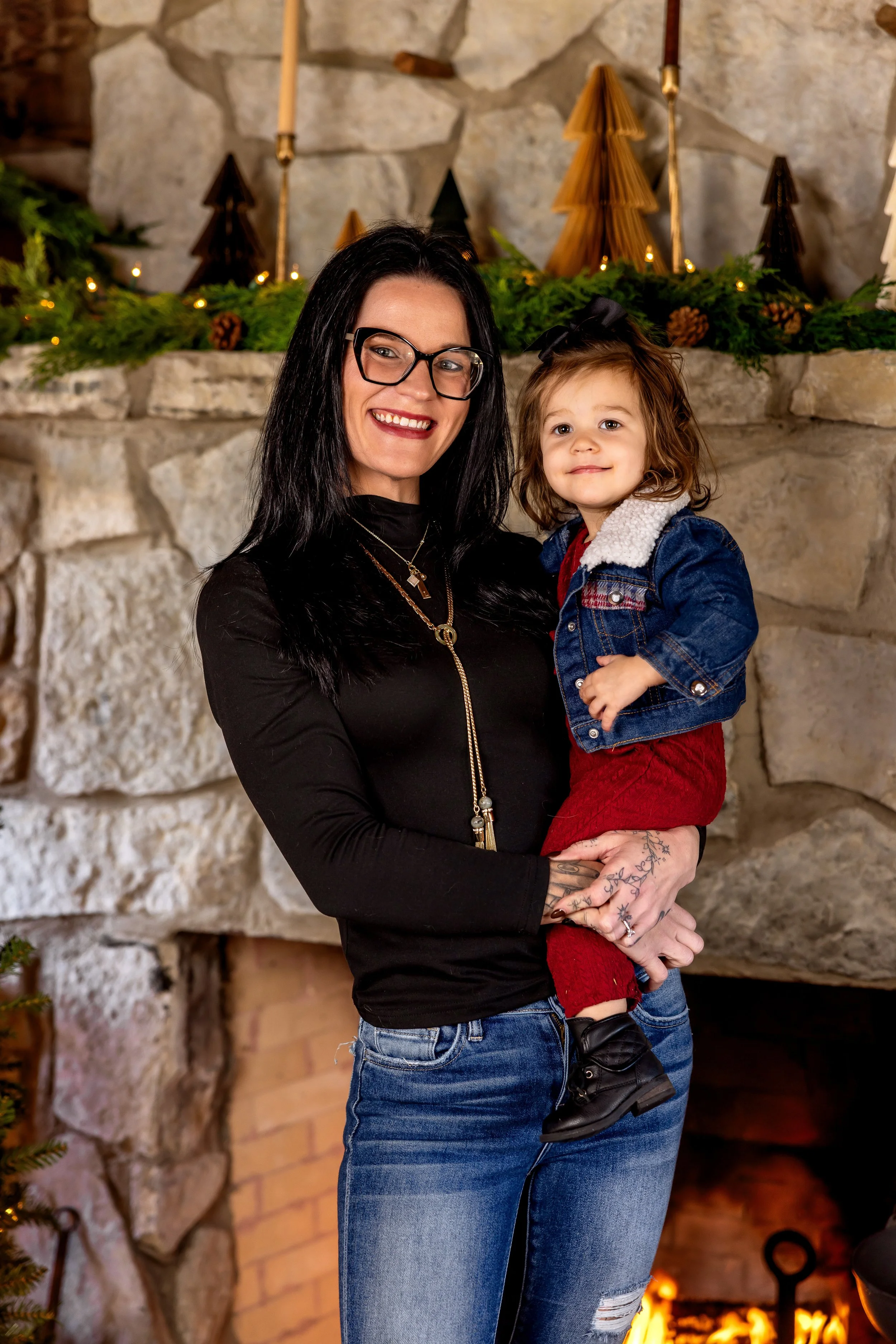 A woman with black hair, glasses, and tattoos, smiling and holding a young girl in front of a decorated fireplace with greenery and candles, during the holiday season.
