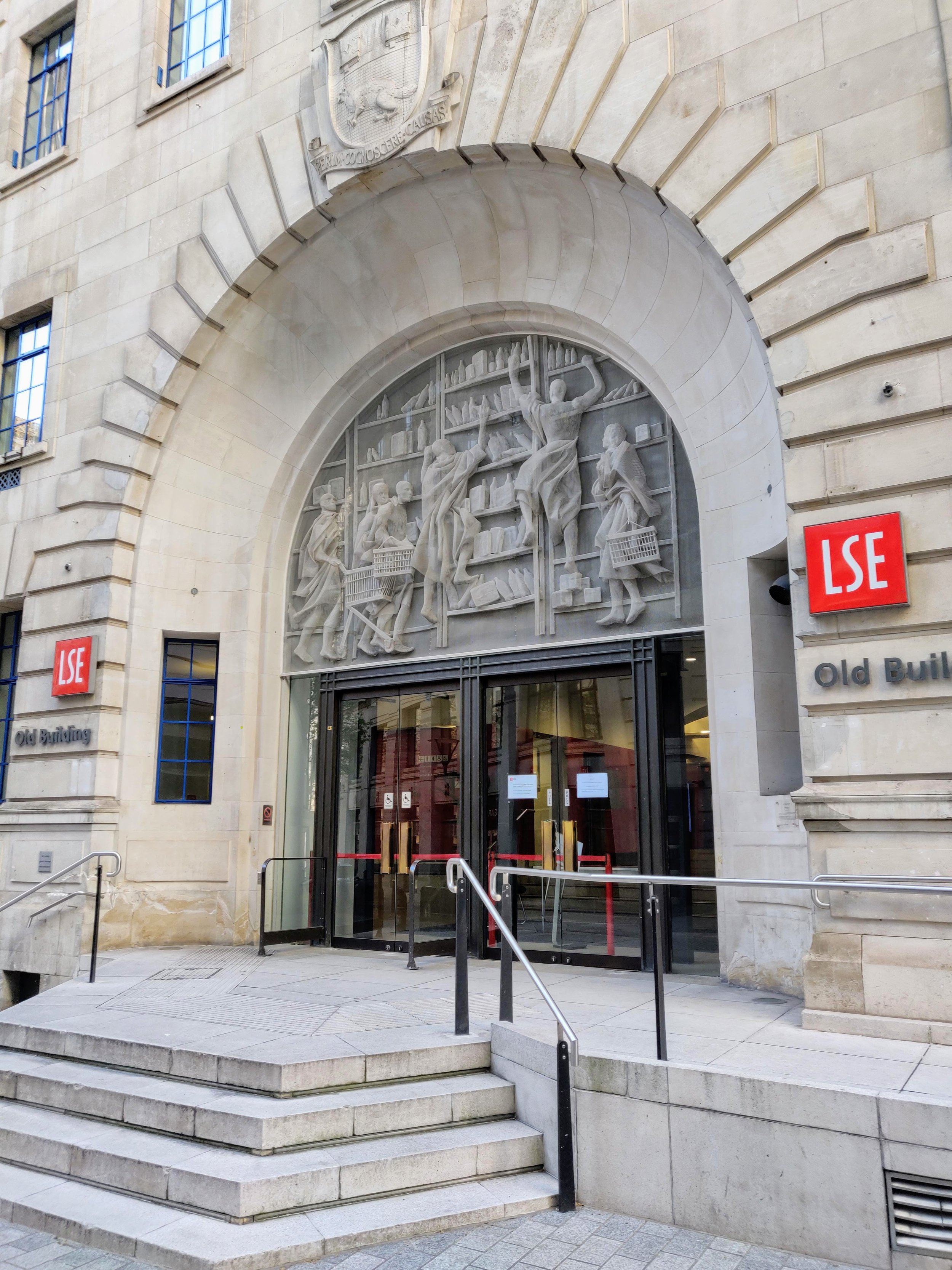 Facade of the London School of Economics (LSE) building with a stone relief sculpture above the entrance depicting books and people in a scholarly scene. Red LSE signs on either side of the entrance, stone stairs leading up to glass doors.