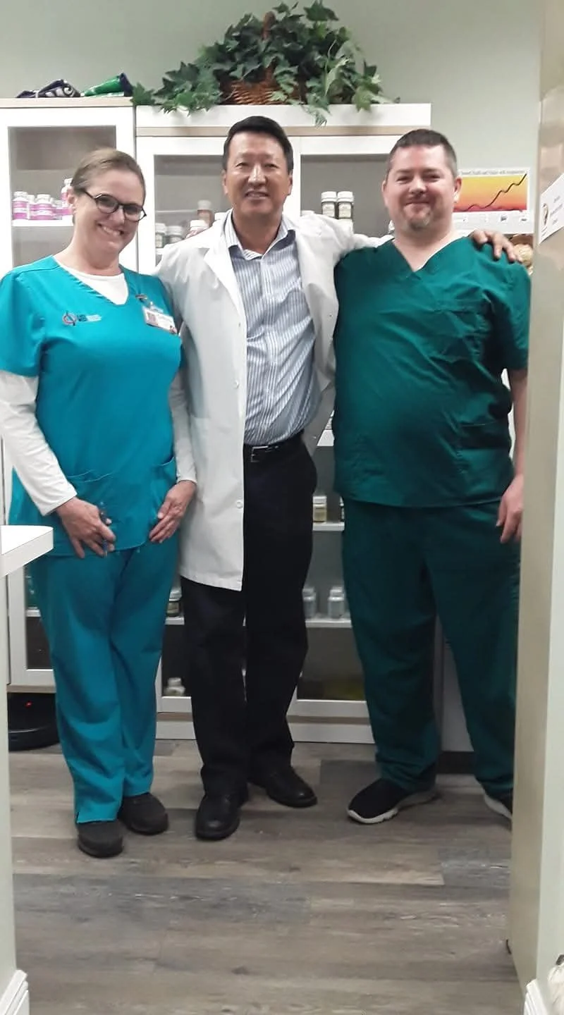 Three healthcare professionals, two men and one woman, standing together in a medical office, smiling at the camera, with a cabinet and plants in the background.