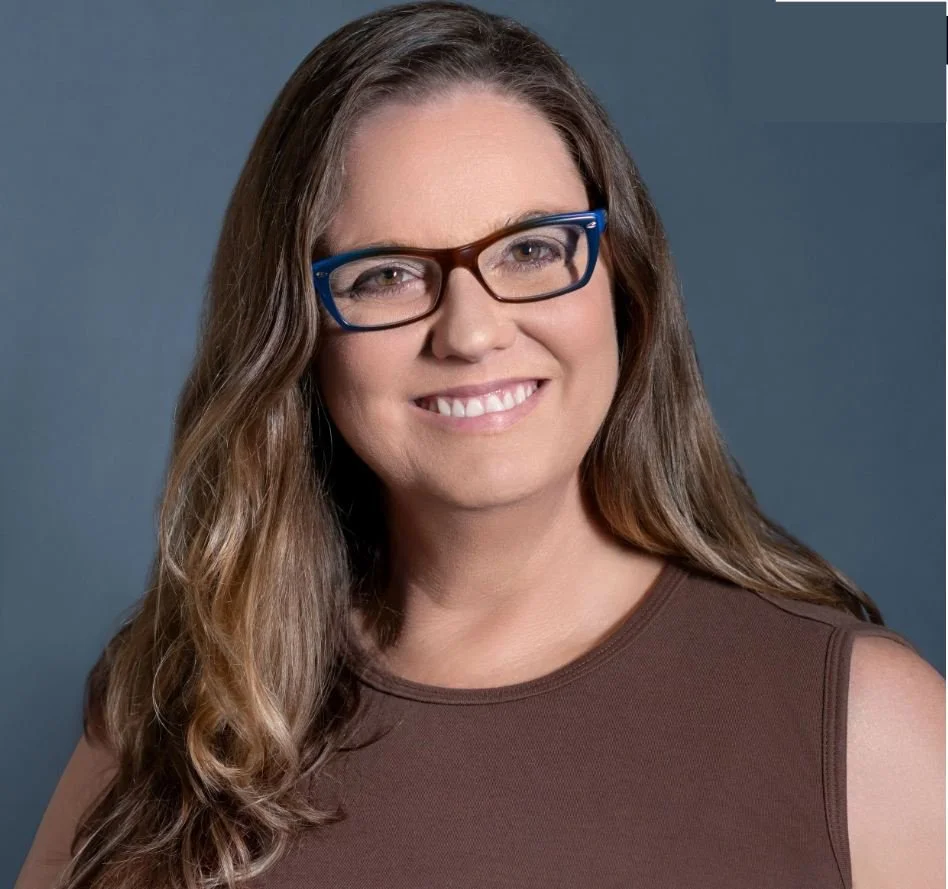 A woman with long brown hair wearing black and blue glasses and a sleeveless brown top, smiling against a dark gray background.