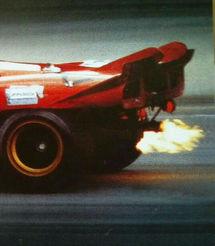 Close-up of a race car's rear end with visible exhaust flames and a racing tire, on a track at night.