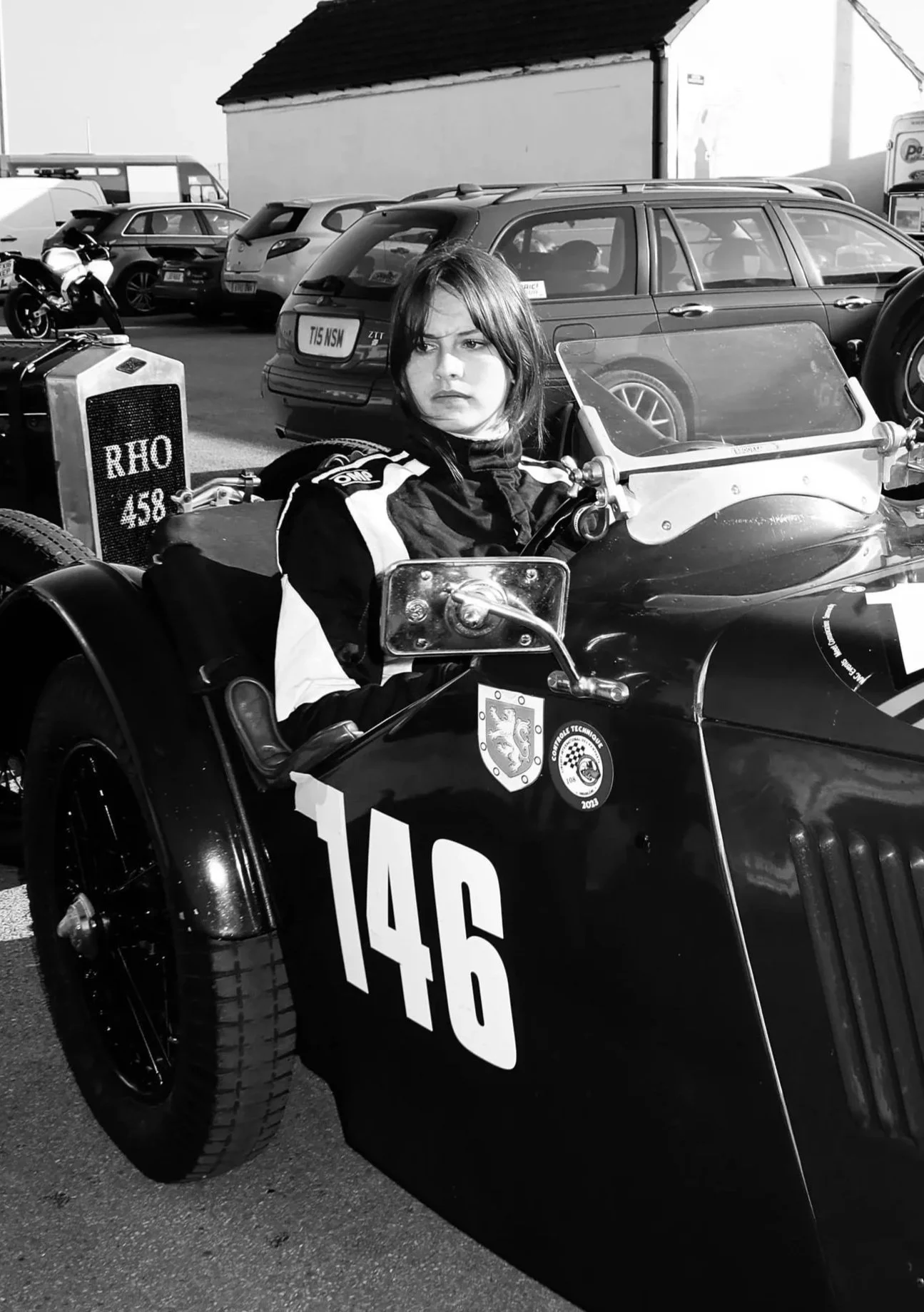 A young woman with dark hair sitting in a vintage race car with the number 146 on the side. She is dressed in a racing suit, and there are several cars and motorcycles in the background, likely at a racetrack or car event.