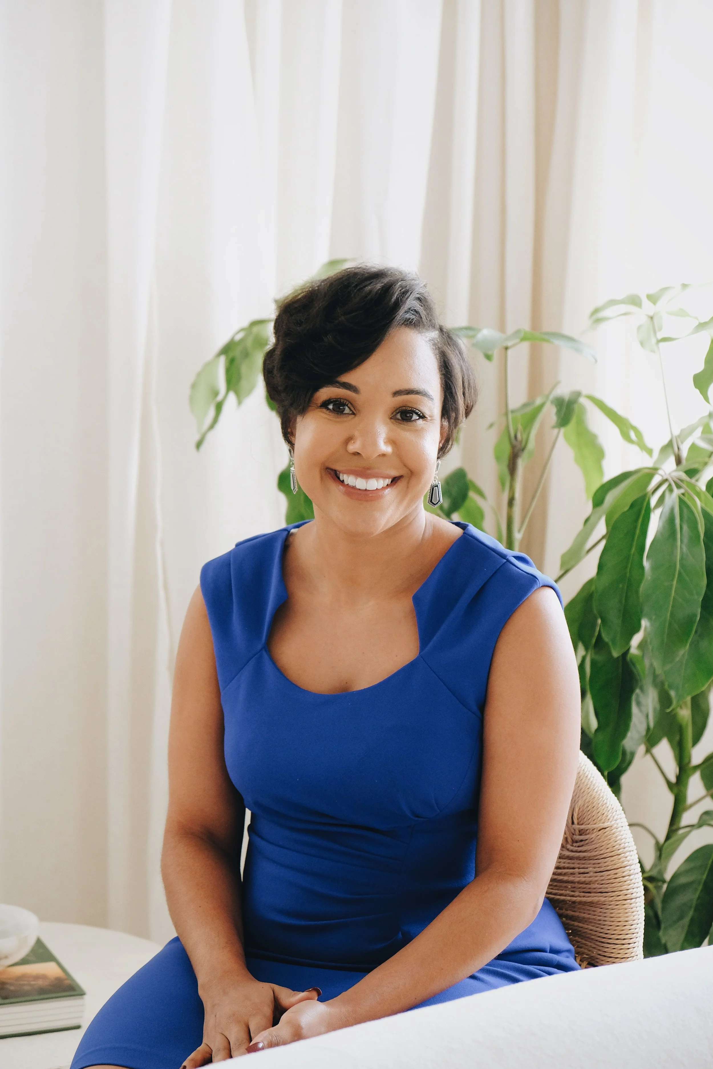A woman with short dark hair and earrings, smiling, wearing a royal blue dress, sitting in a room with white curtains and a large green plant behind her.