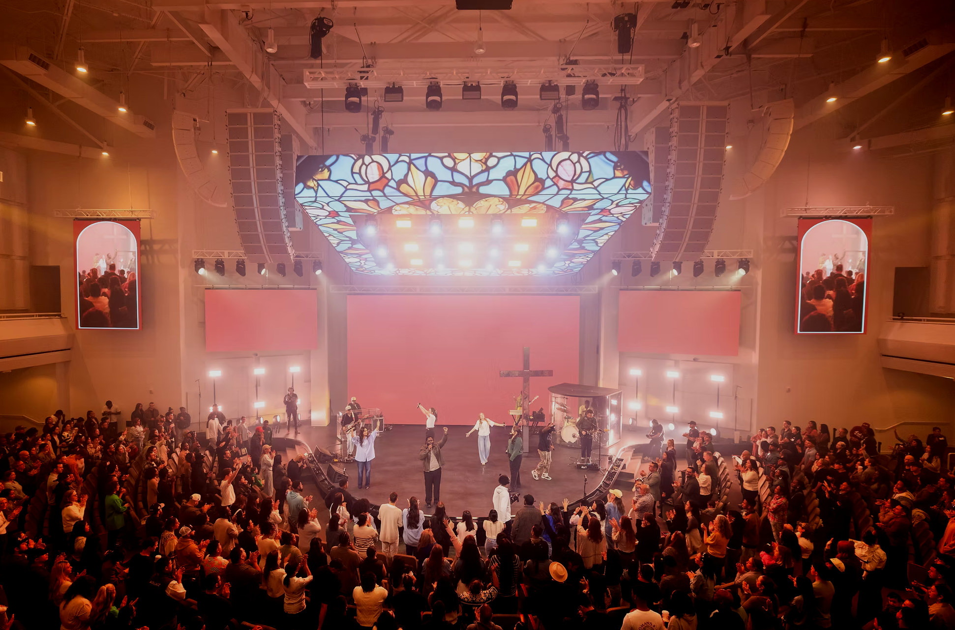 A large indoor church service or concert with a stage, musicians, and a congregation raising their hands. The stage features a stained-glass ceiling design, a cross, and electronic screens displaying visuals. The atmosphere is lively with warm lighting, and large screens on either side display images of the event.
