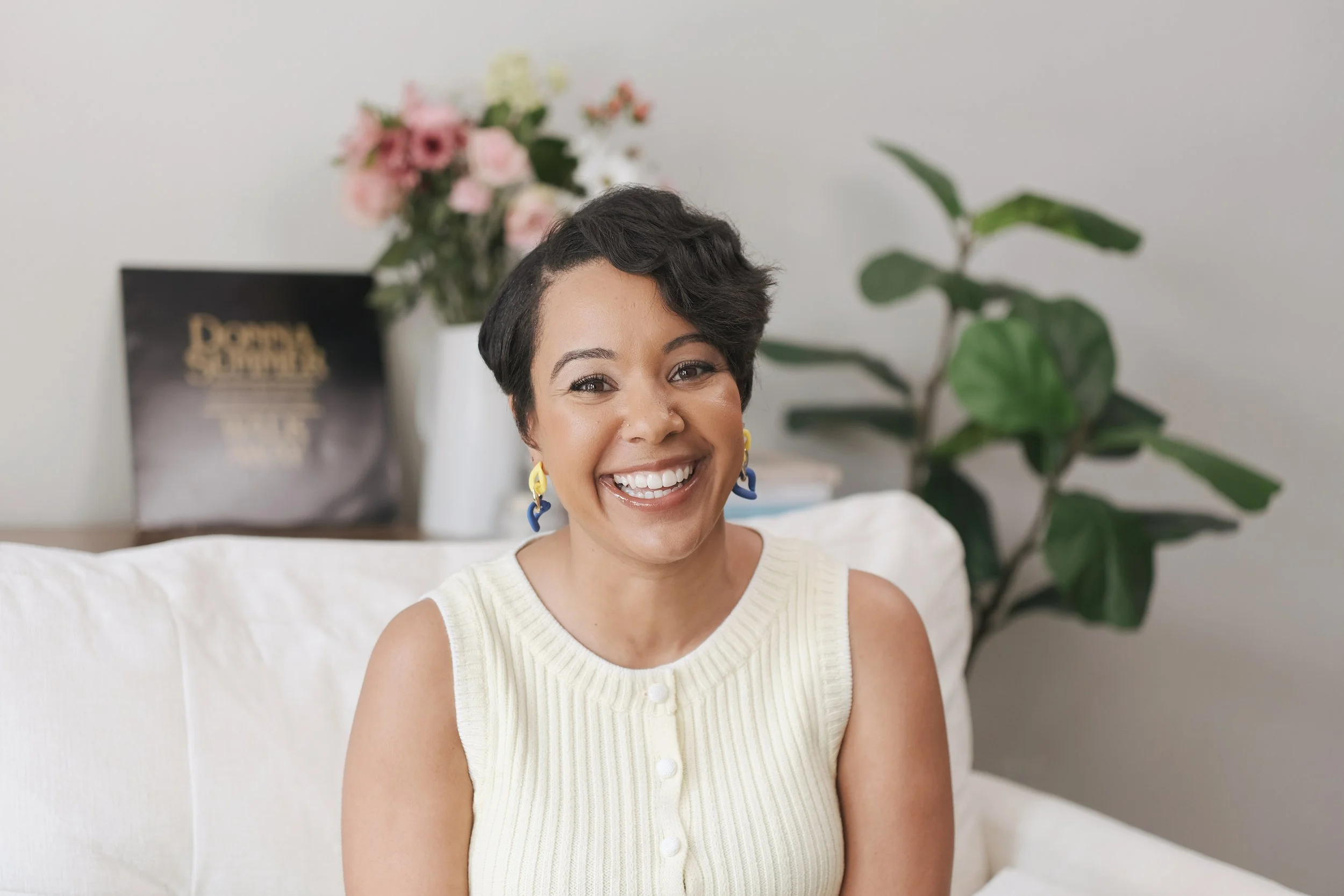A woman with short dark hair, smiling, wearing a cream-colored sleeveless cardigan, sitting on a white sofa with a background of a plant and a flower arrangement.