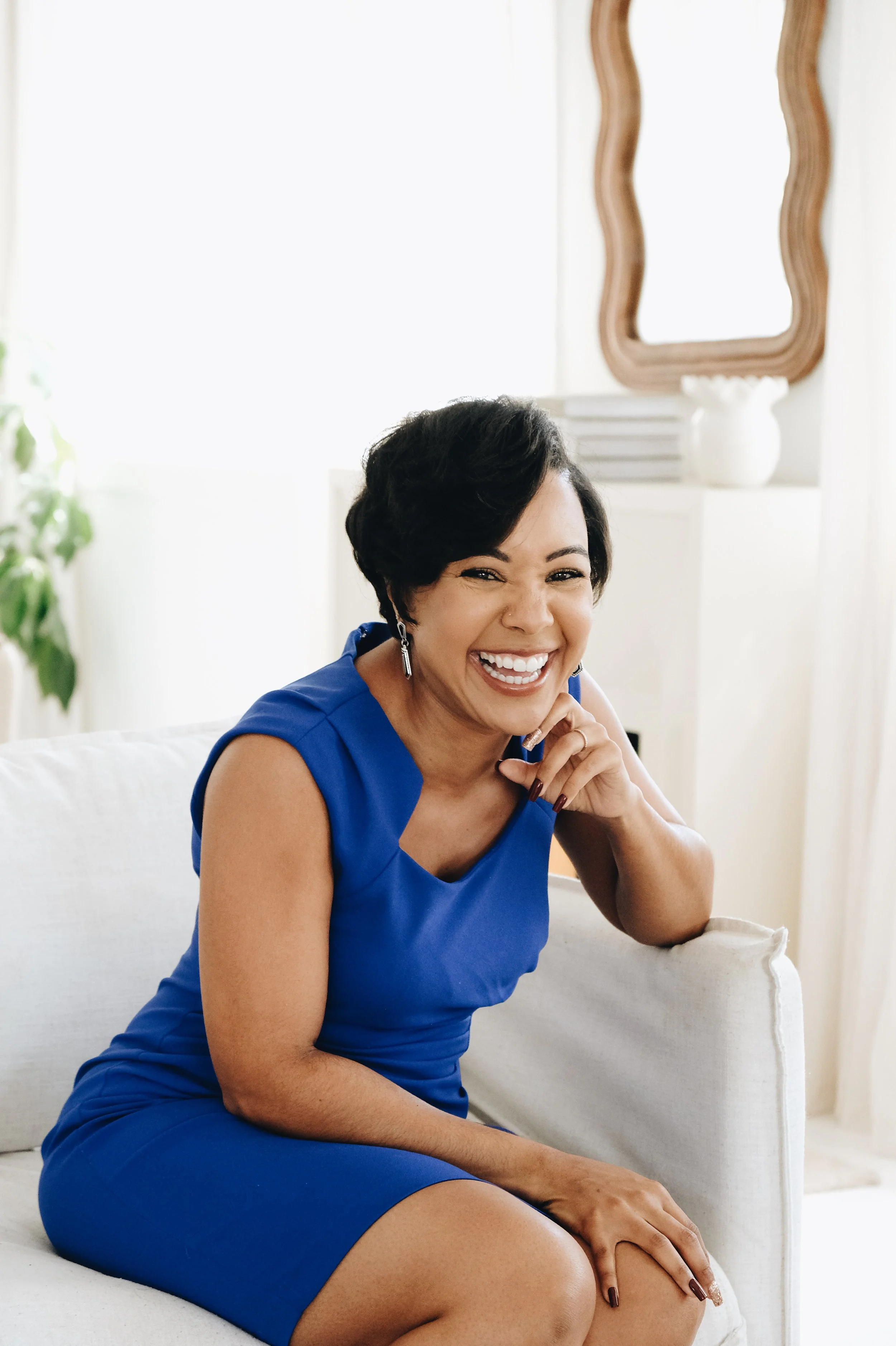 A woman wearing a blue dress sitting on a white sofa, smiling broadly and laughing in a well-lit room with a mirror and plants in the background.