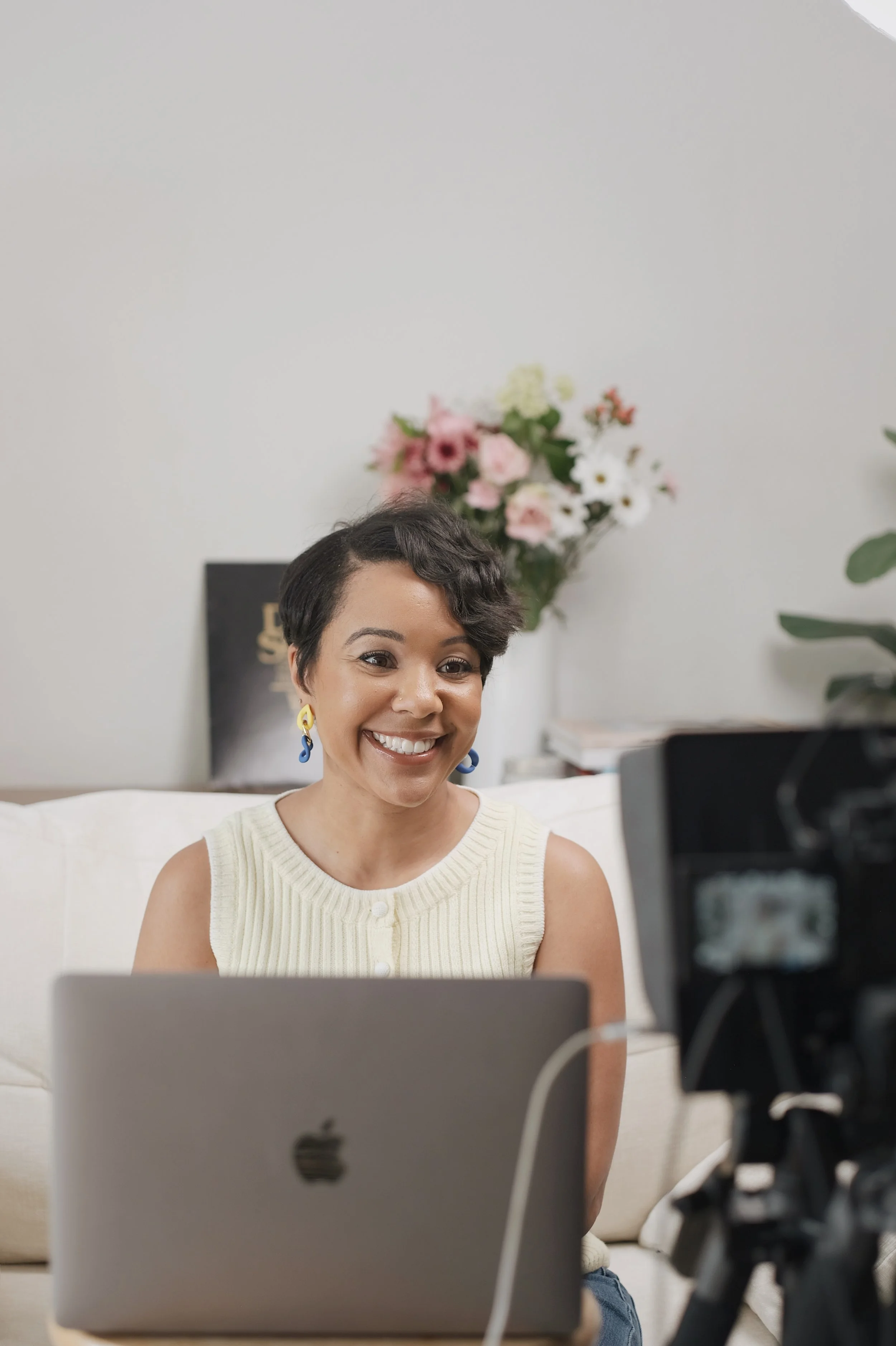 A woman with short dark hair, wearing a sleeveless cream-colored top and colorful earrings, smiling during a video call on her laptop, with a webcam and camera set up in front of her, against a background with flowers and books.