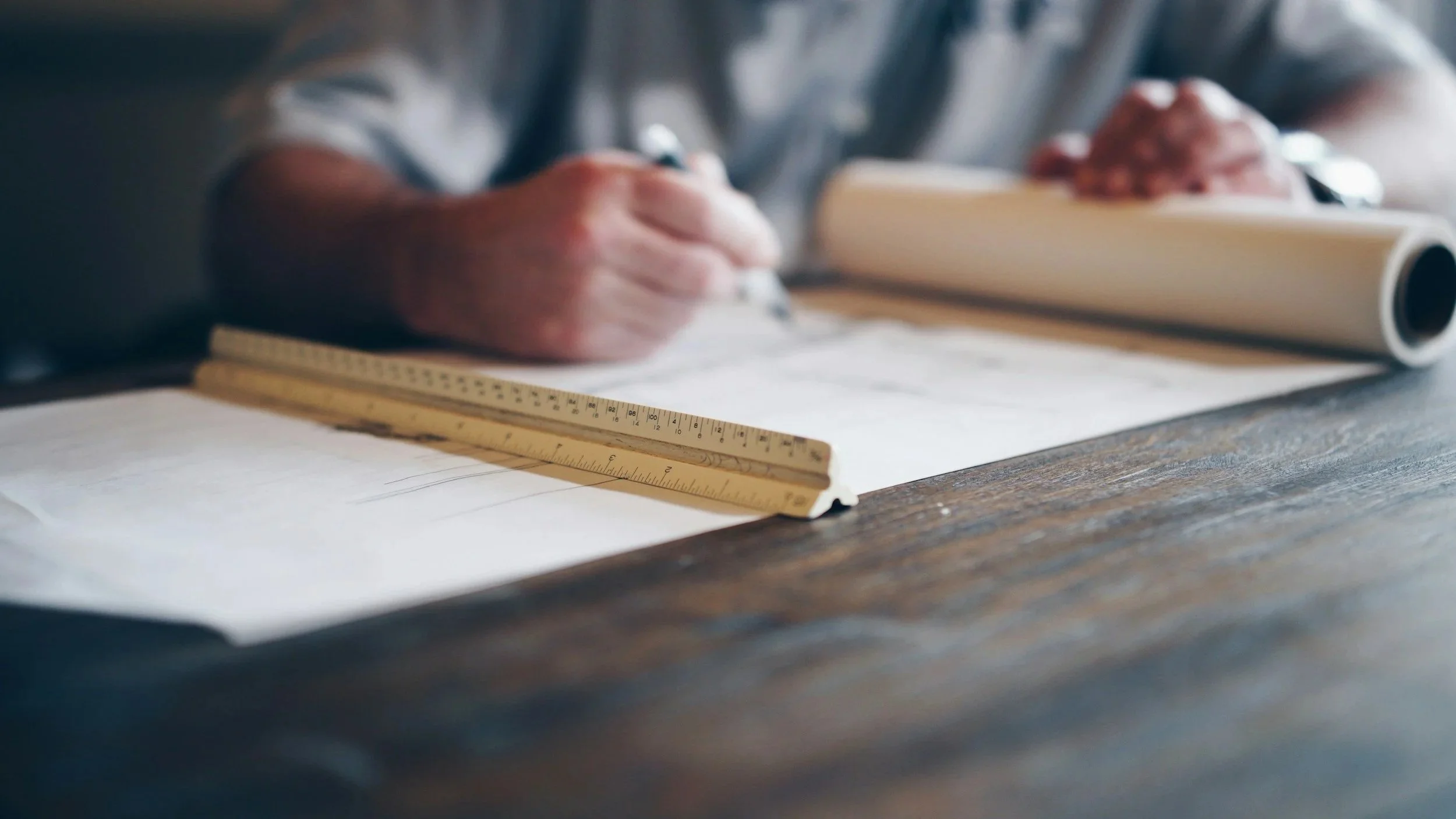 Person working on architectural or engineering plans, using a ruler on a wooden table with a rolled-up blueprint in view.