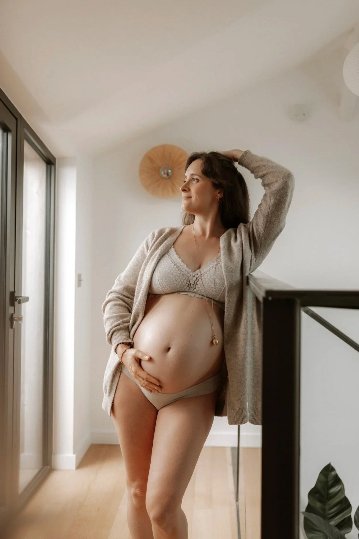 Femme enceinte dans une pièce lumineuse, portant un soutien-gorge beige, un cardigan gris clair, et tenant son ventre avec une main.