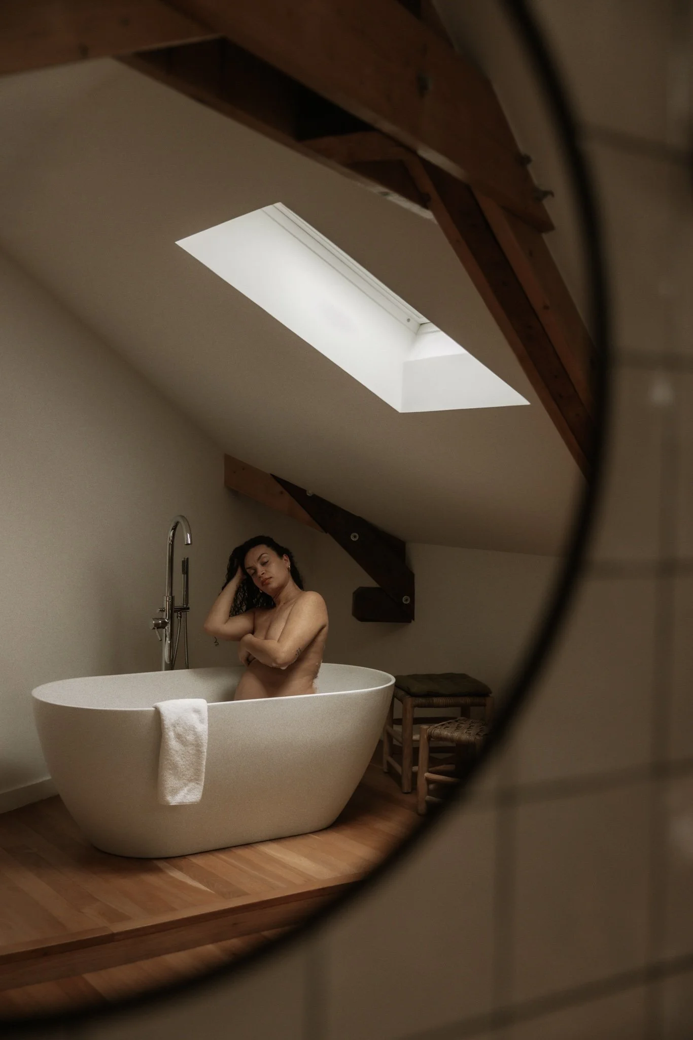 Jeune femme nue assise dans une baignoire blanche, regardant vers le bas, vue à travers un miroir, dans une pièce avec plafond en bois et fenêtre de lumière naturelle.