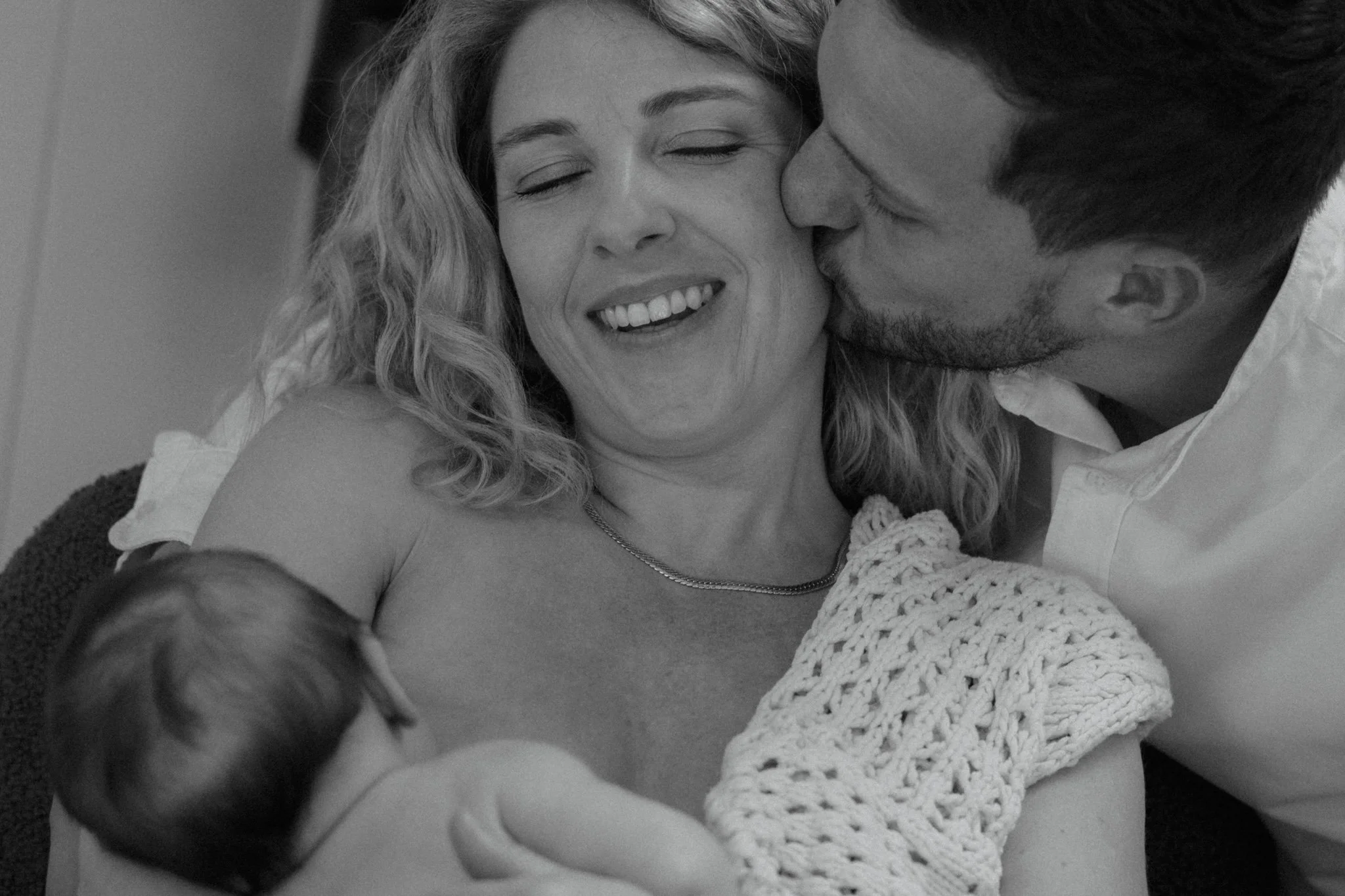 Une famille lors de la naissance d'un bébé, la mère en train d'allaiter, le père embrassant la mère, tous souriants.