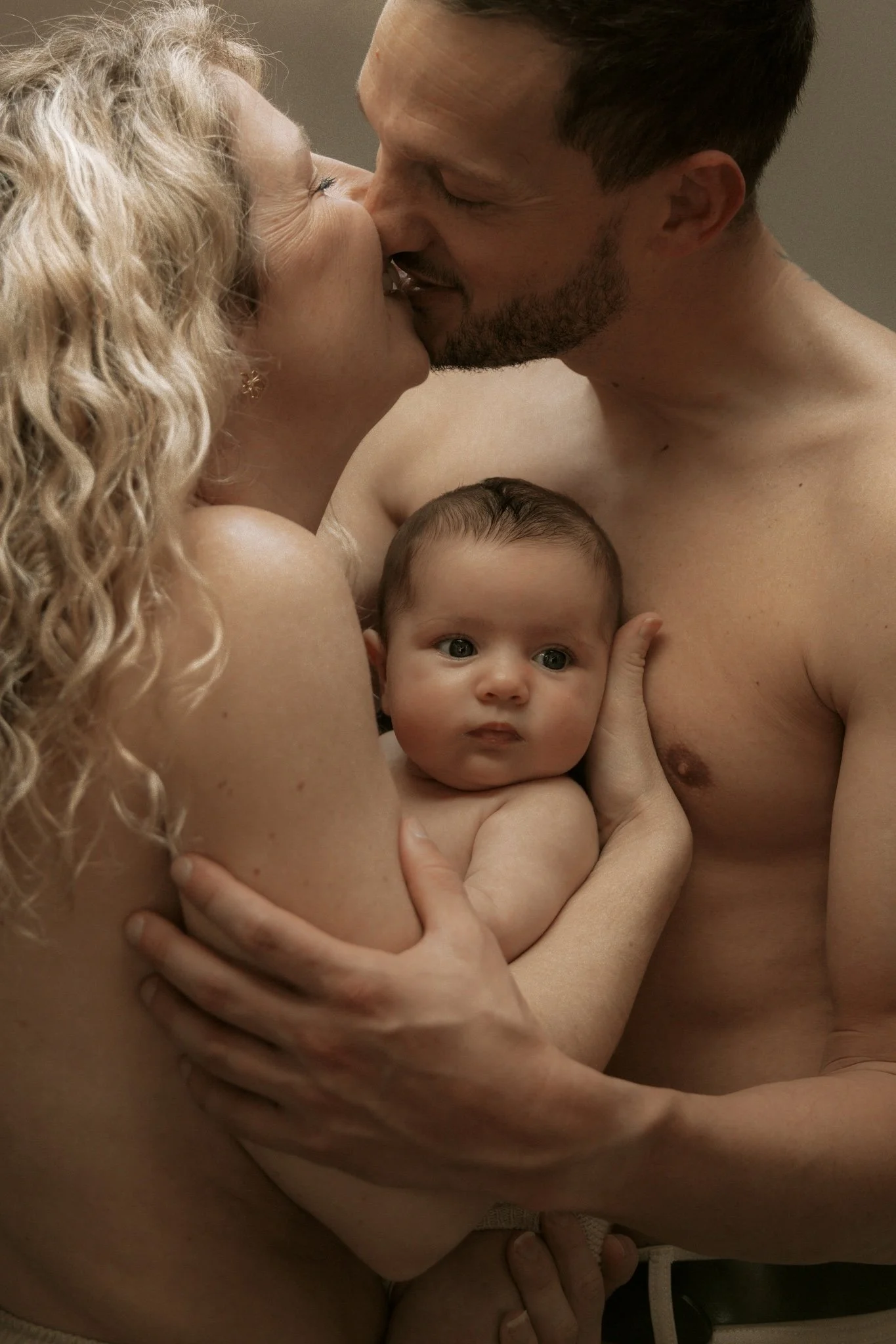 Un couple de parents avec leur bébé, échangeant un baiser tendrement, le bébé regarde simplement.