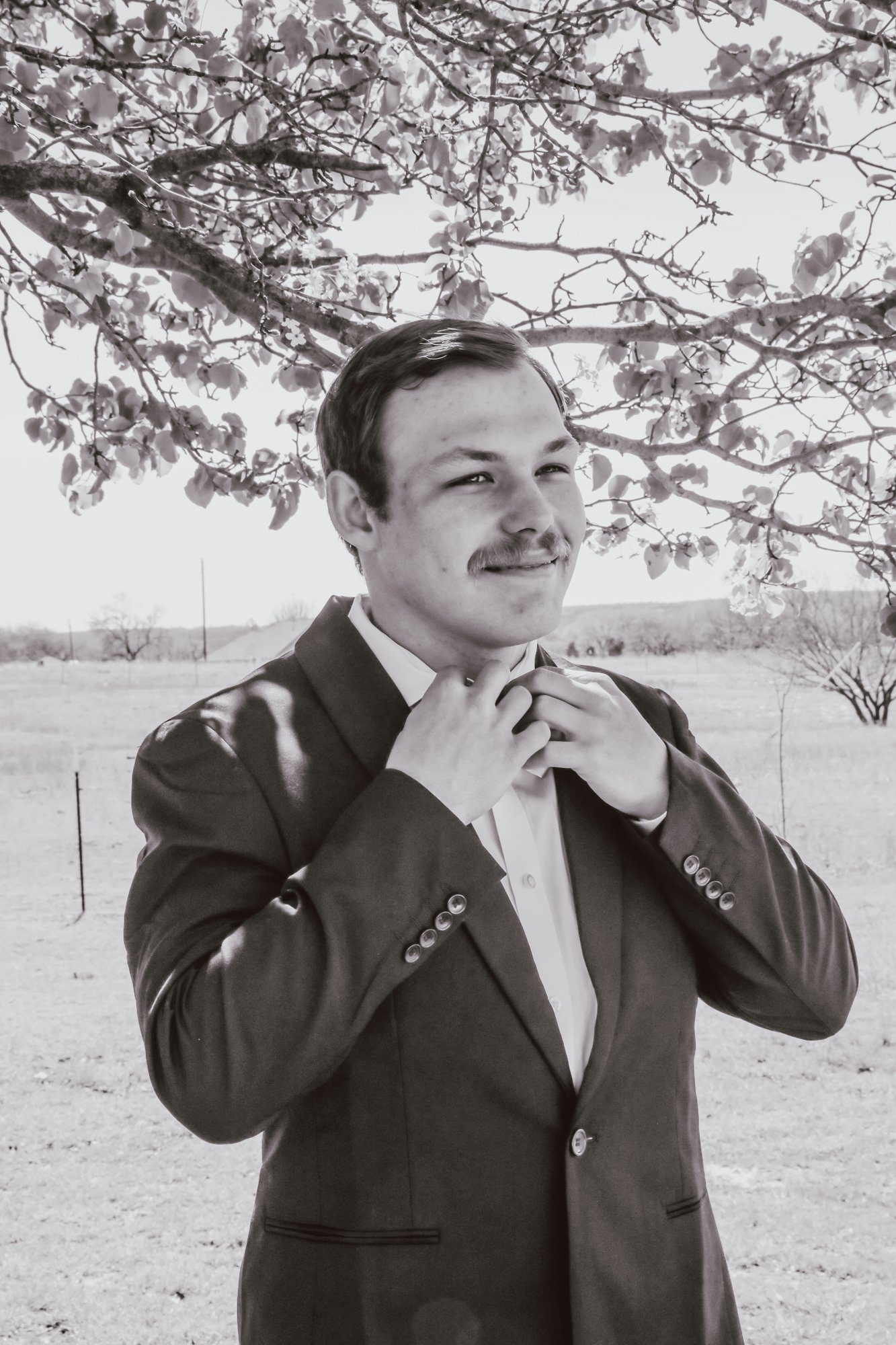 A man adjusting his bow tie outdoors in front of a tree with blossoms, wearing a suit.