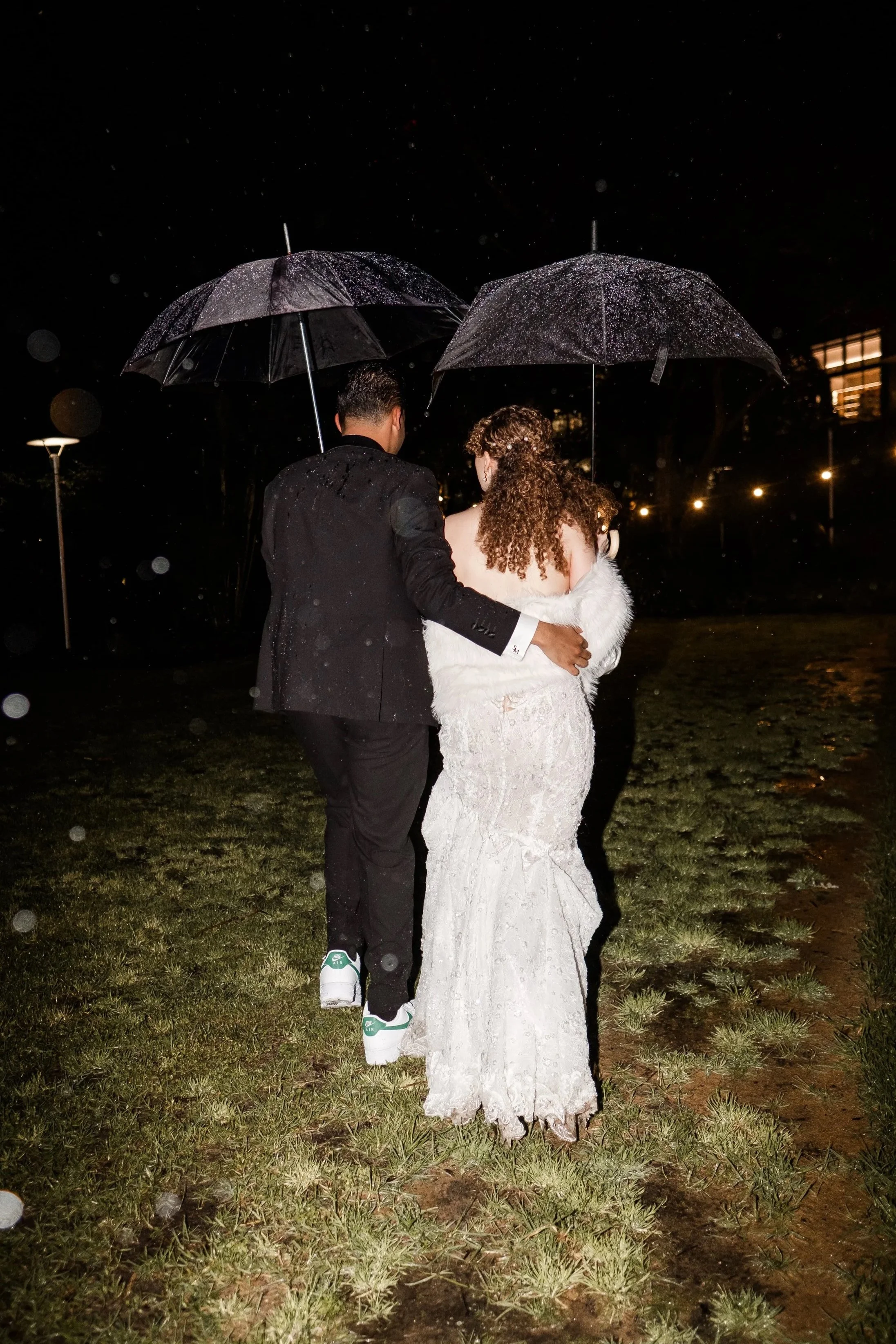 A couple walking outdoors at night in the rain, holding umbrellas, with the woman in a white dress and the man in a black suit.