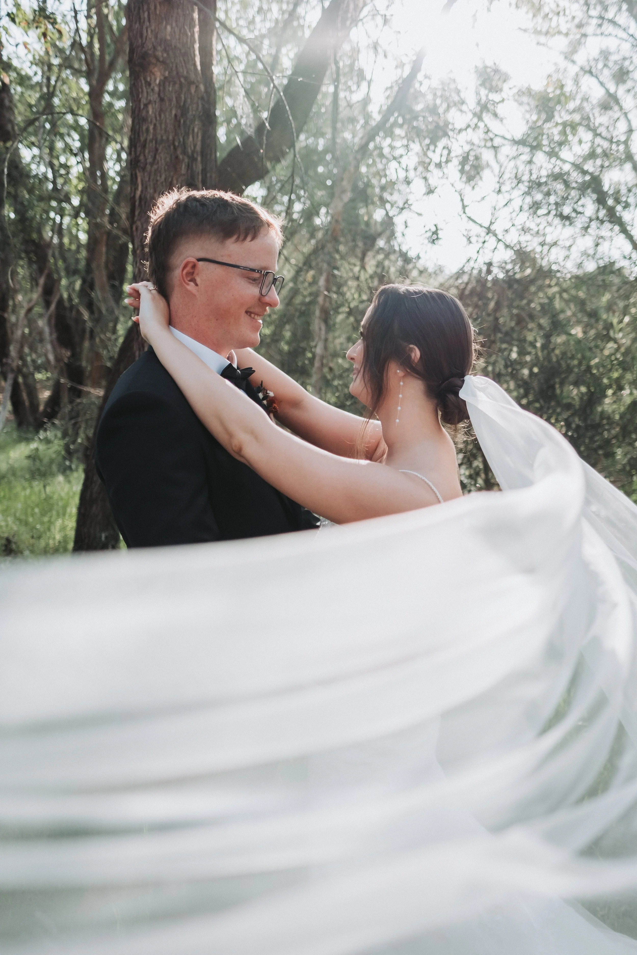 A bride and groom on their wedding day in an outdoor setting, embracing each other with trees and sunlight in the background.