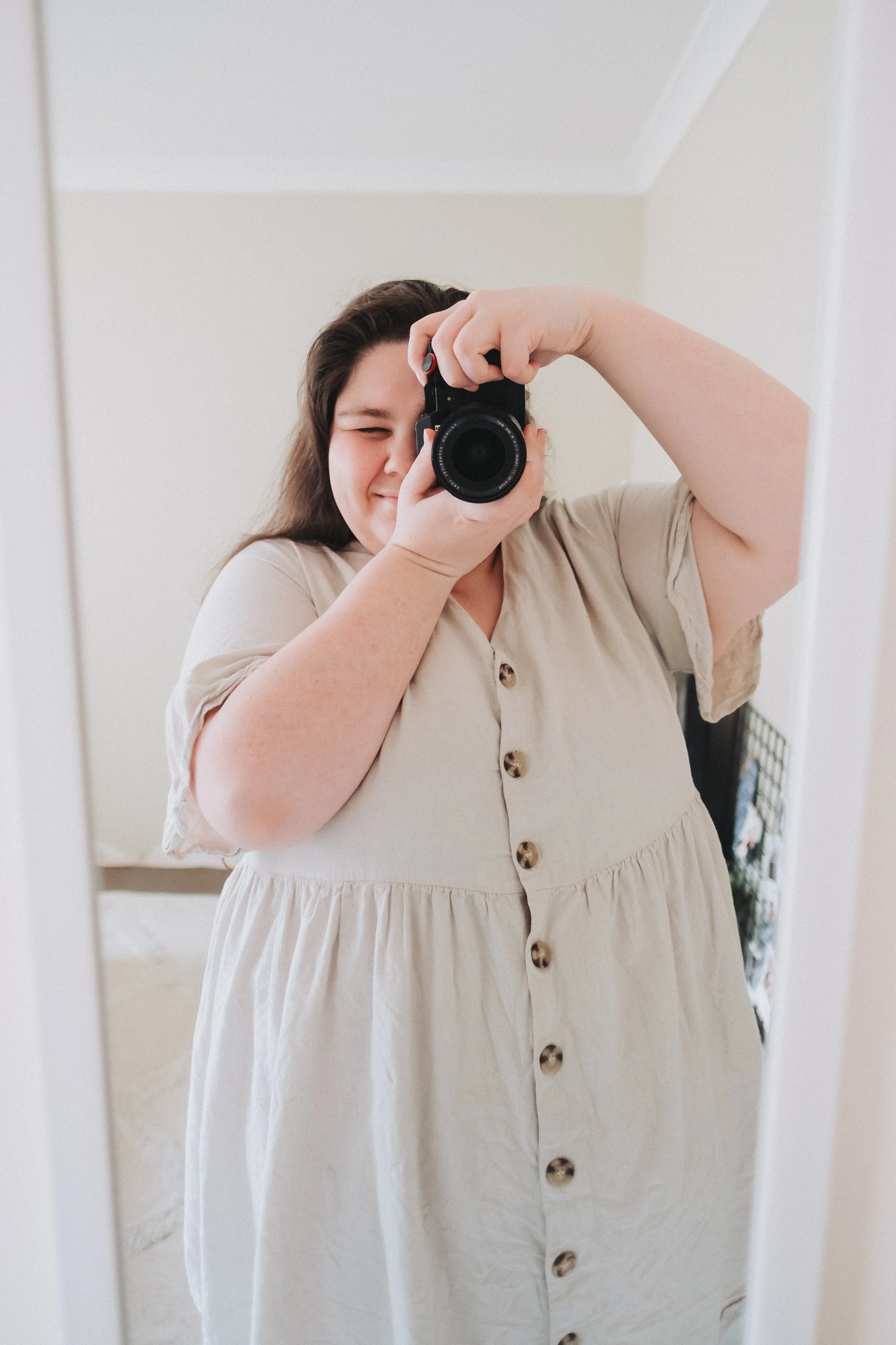 A woman taking a selfie in a mirror with a camera, smiling, wearing a beige dress with buttons, in a bright room.