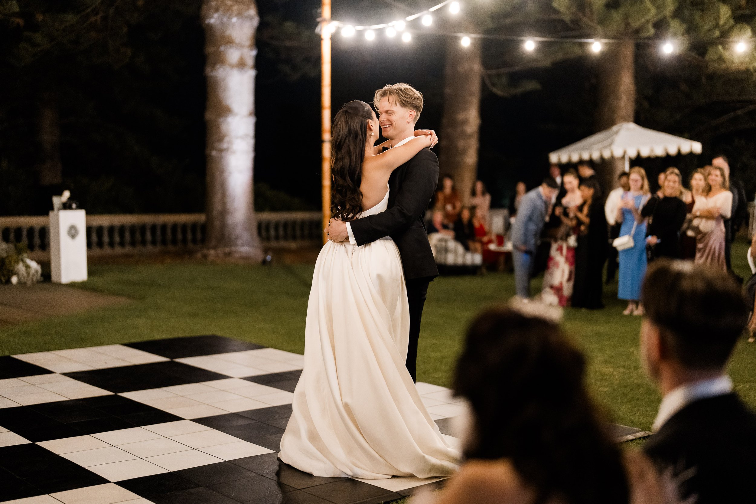 A bride and groom are dancing together outdoors at night under string lights, surrounded by guests.