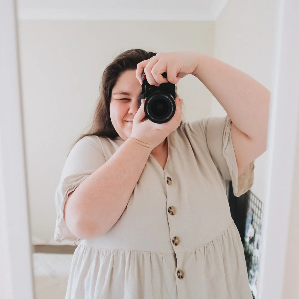 Person taking a mirror selfie with a camera, making a playful face, wearing a beige button-up dress