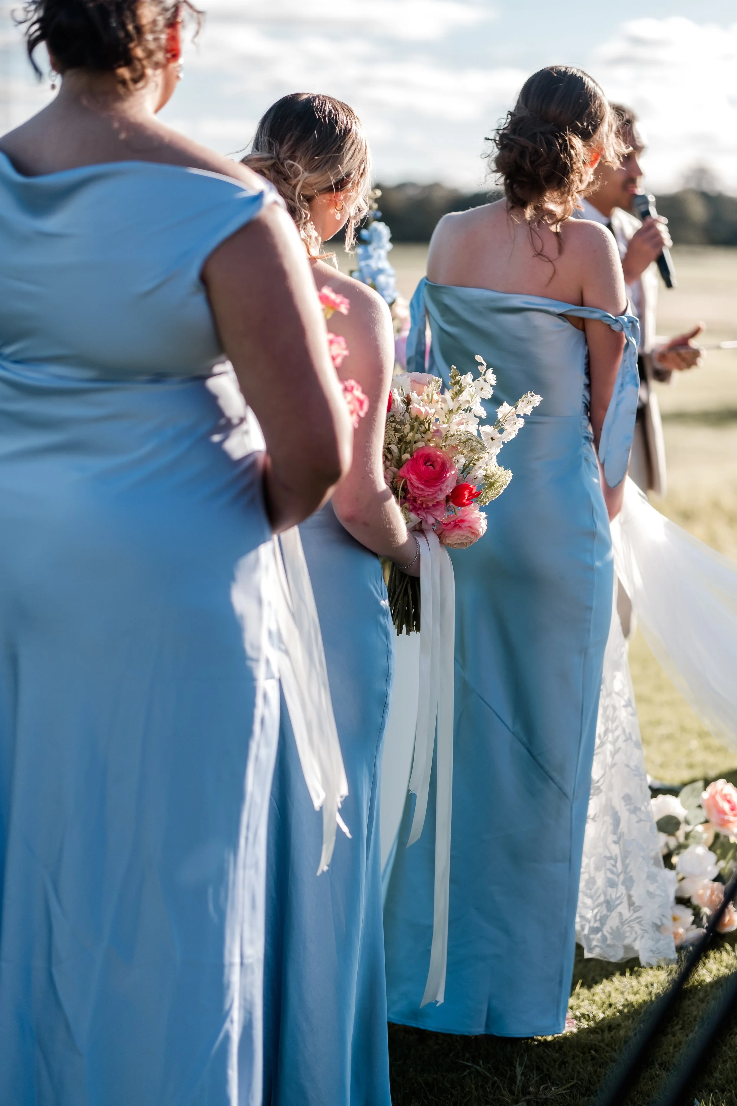 Four women wearing light blue dresses standing outdoors during a wedding ceremony, holding a bouquet of flowers. A man is speaking into a microphone in the background.
