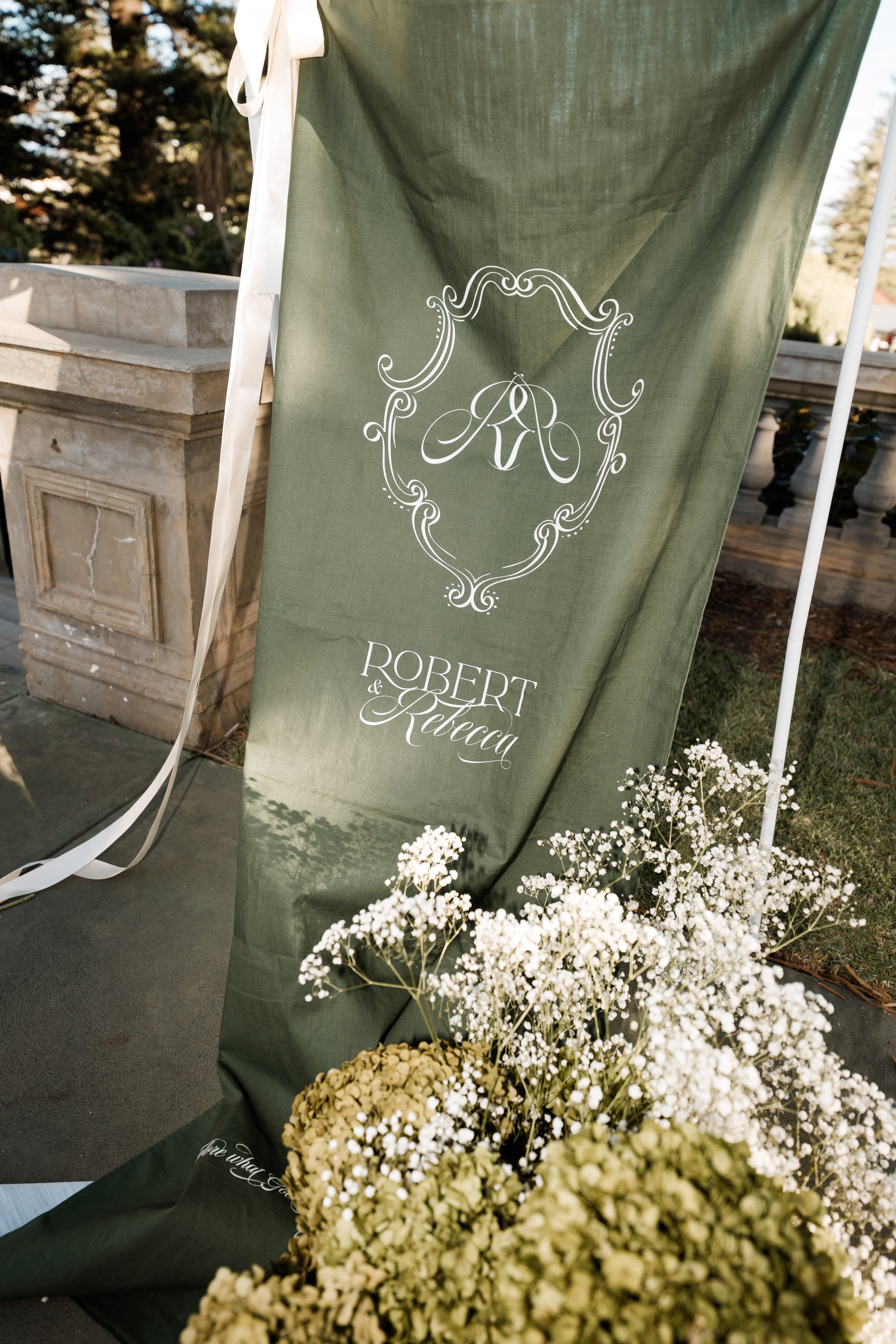 Green fabric banner with white ornate monogram and the names Robert and Rebecca surrounded by white flowers outside.