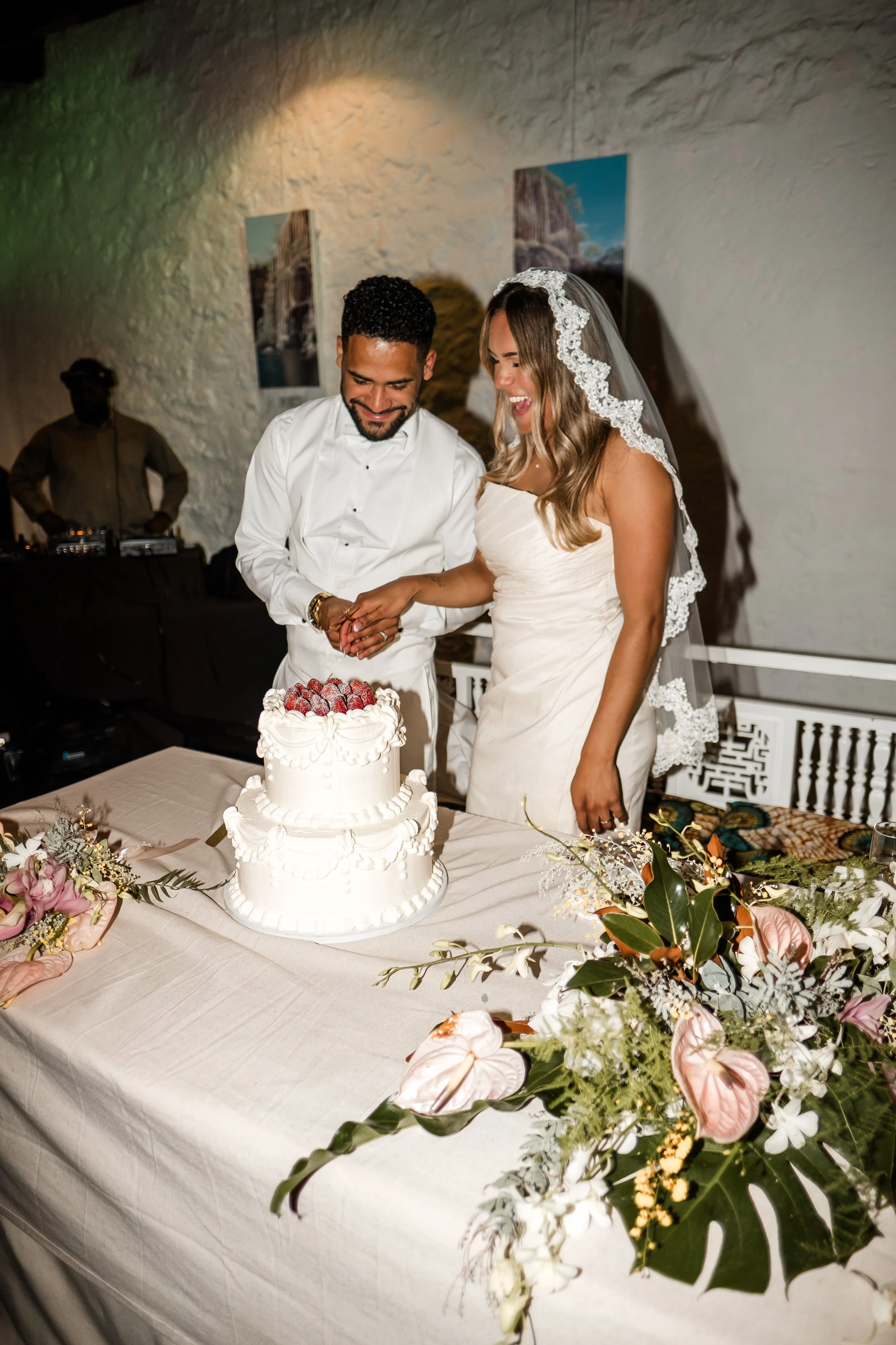 Bride and groom cutting a wedding cake together, smiling, with wedding decorations and floral arrangements on the table.