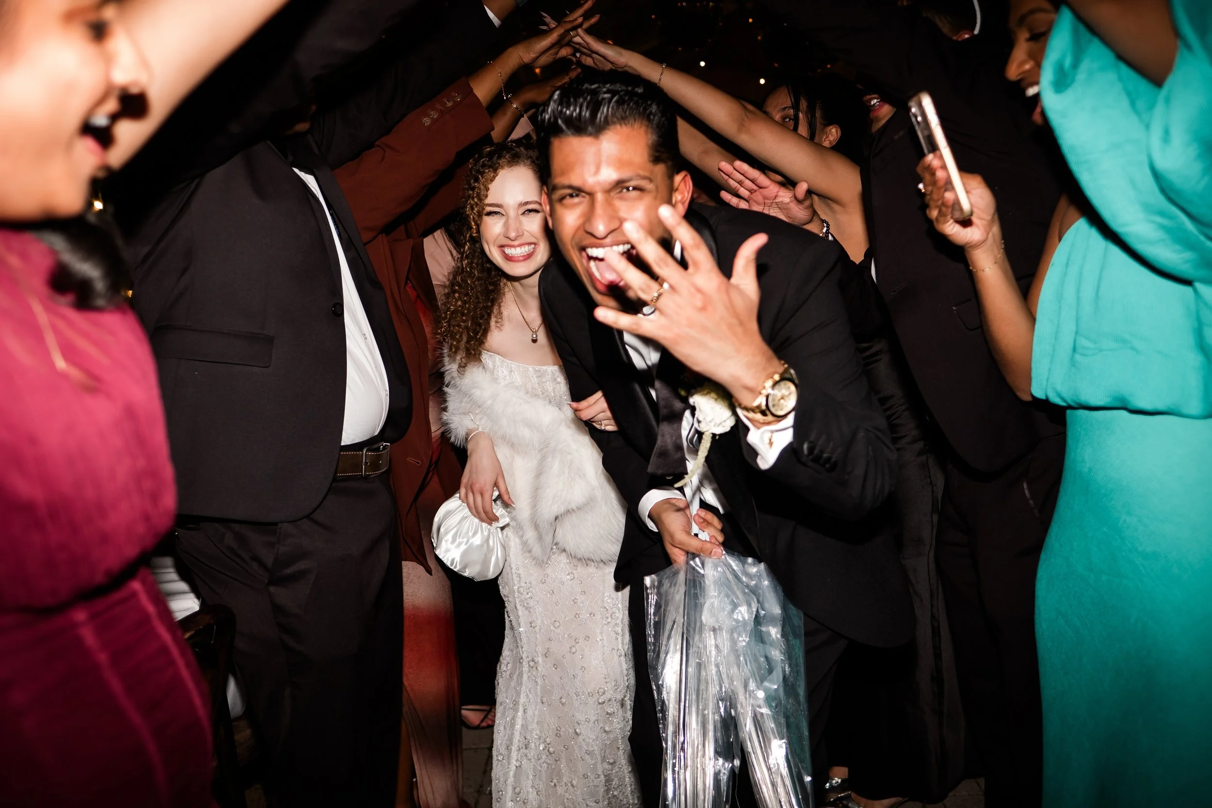Group of people celebrating at a wedding reception, with a man in a black tuxedo and a woman in a white wedding dress at the center, smiling and having fun.