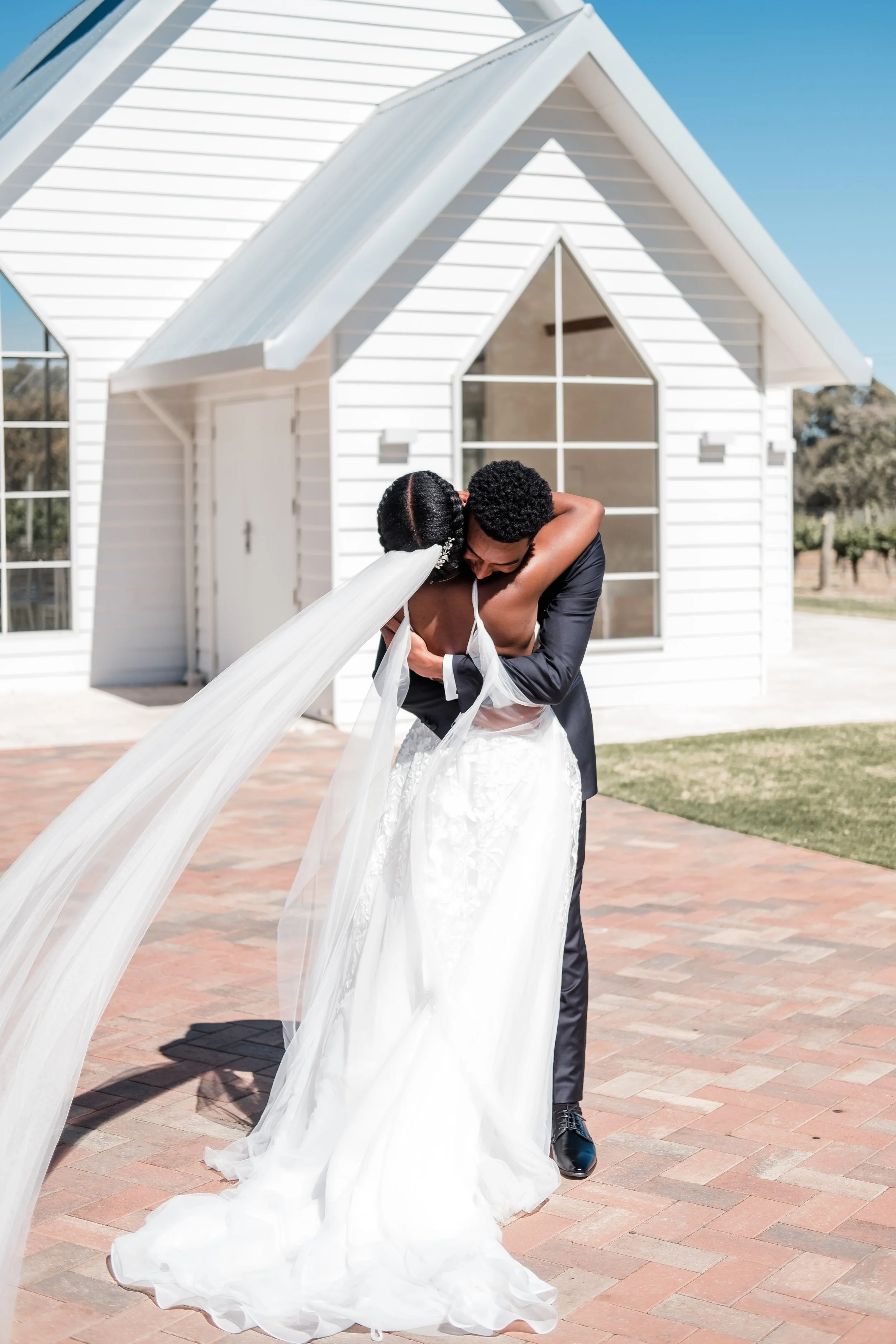 A bride and groom sharing a dance outside a white house on a sunny day. The bride wears a wedding dress with a long veil, and the groom is in a black suit. They are embracing each other closely.