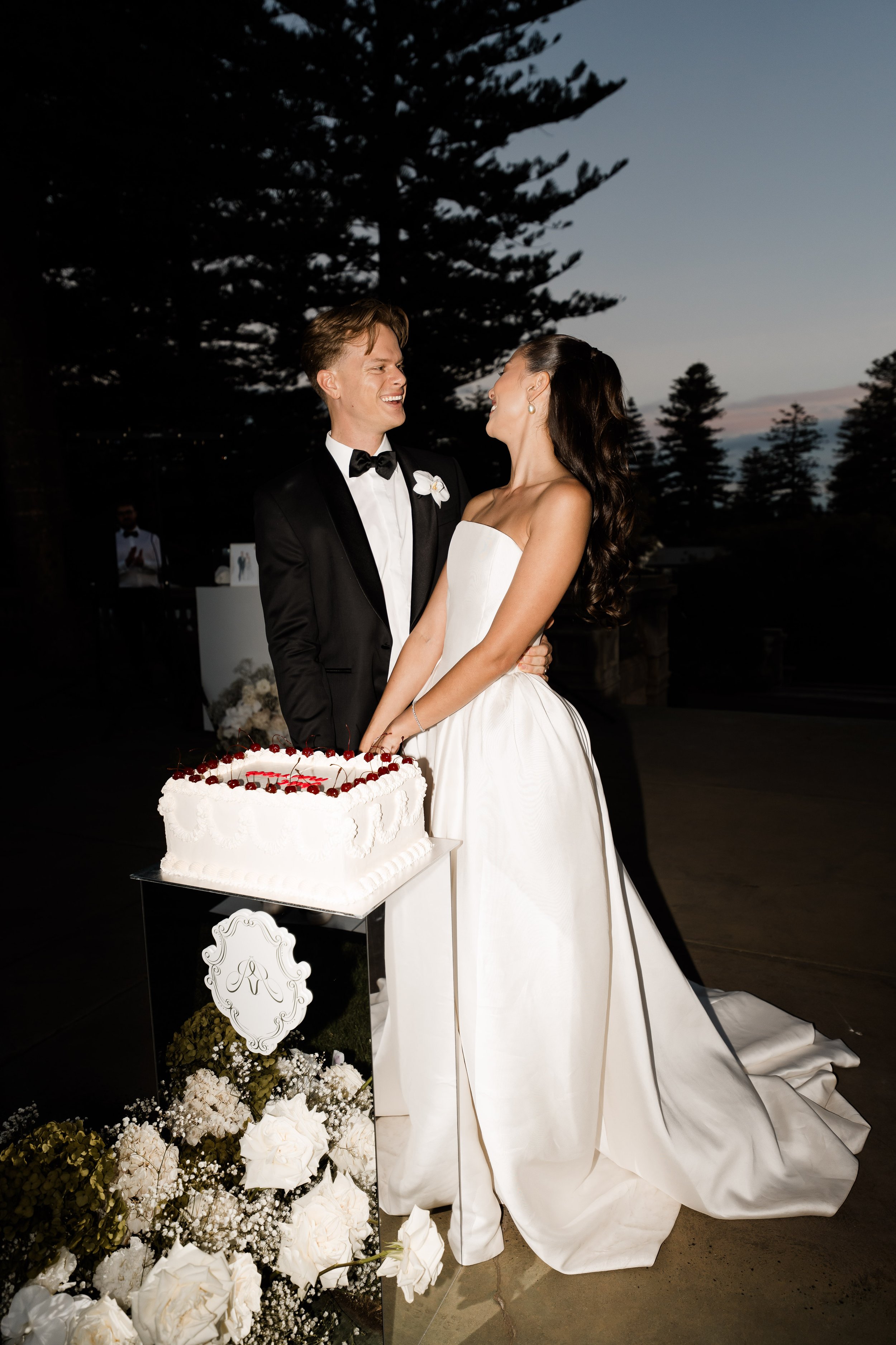 A bride and groom share a moment at their wedding reception, standing next to a wedding cake decorated with cherries and white frosting, outdoors during dusk with trees in the background.