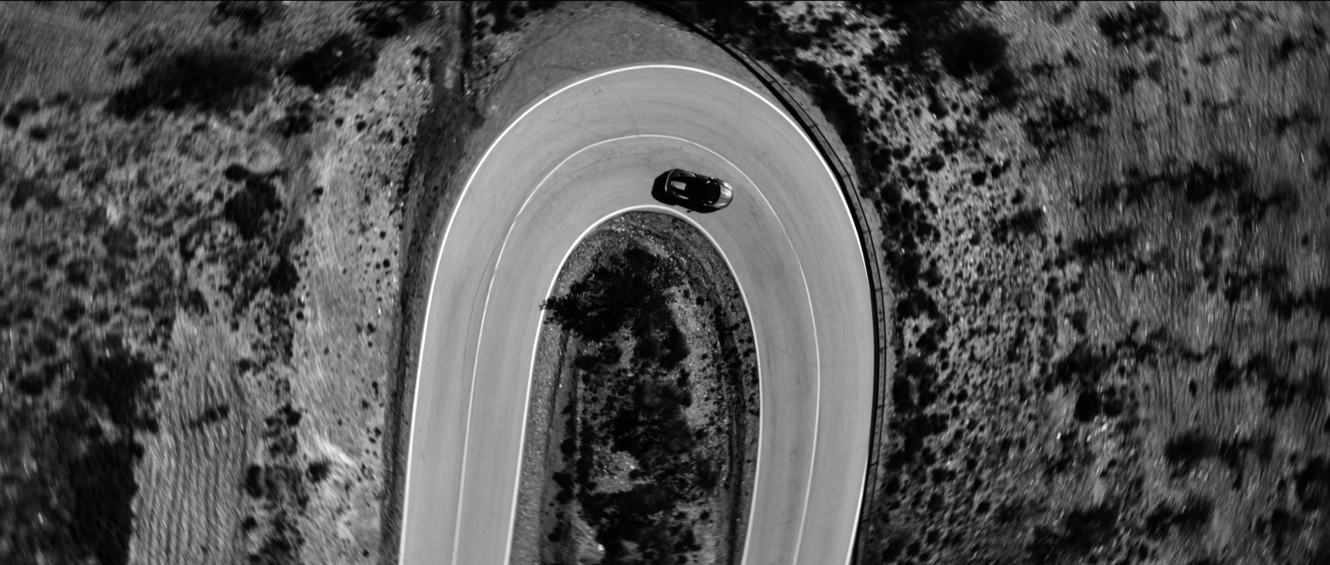 An aerial view of a black car driving on a winding mountain road surrounded by rocky and sparse desert landscape.