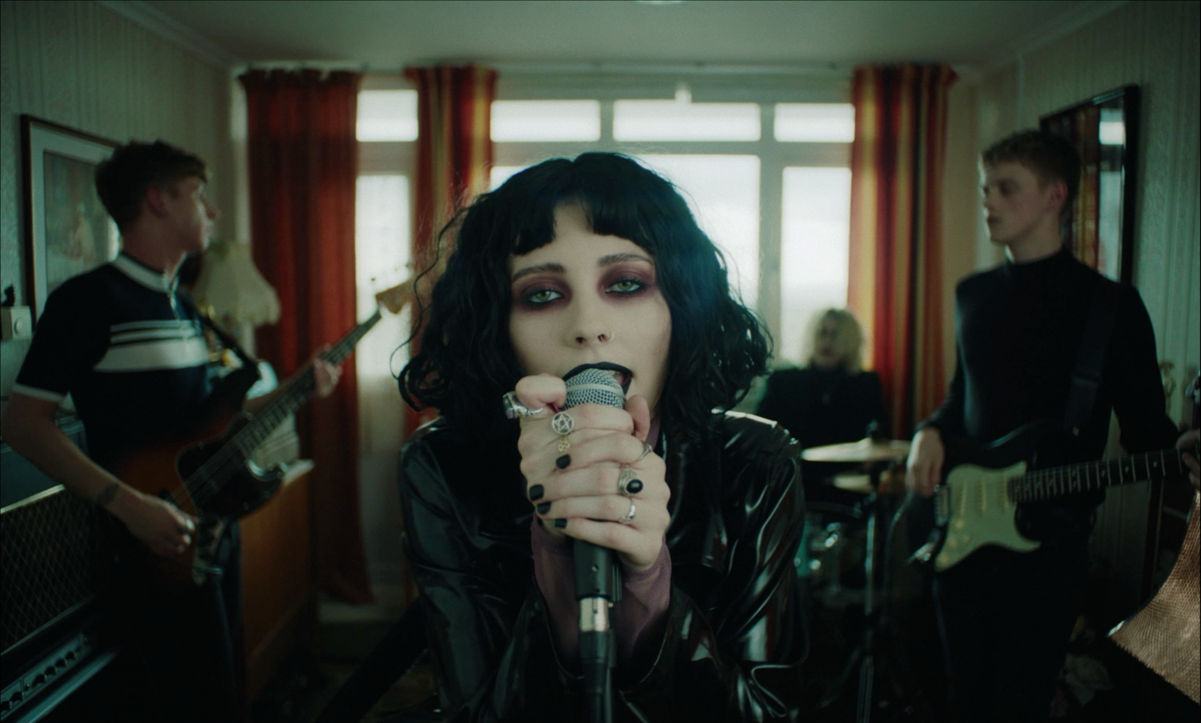 A woman with black, curly hair and dark makeup singing into a microphone in a room with three men playing guitars and drums, with red curtains and large windows in the background.