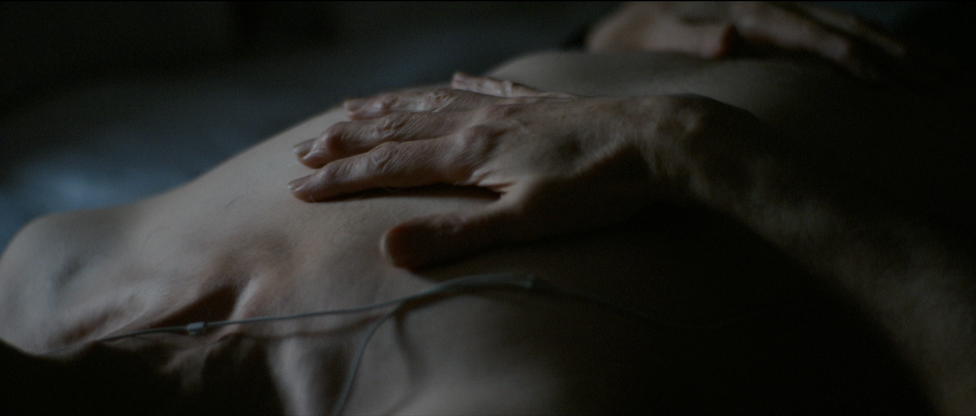 A close-up of a person's hand resting on their bare chest with a medical lead wire attached, in a dimly lit setting.