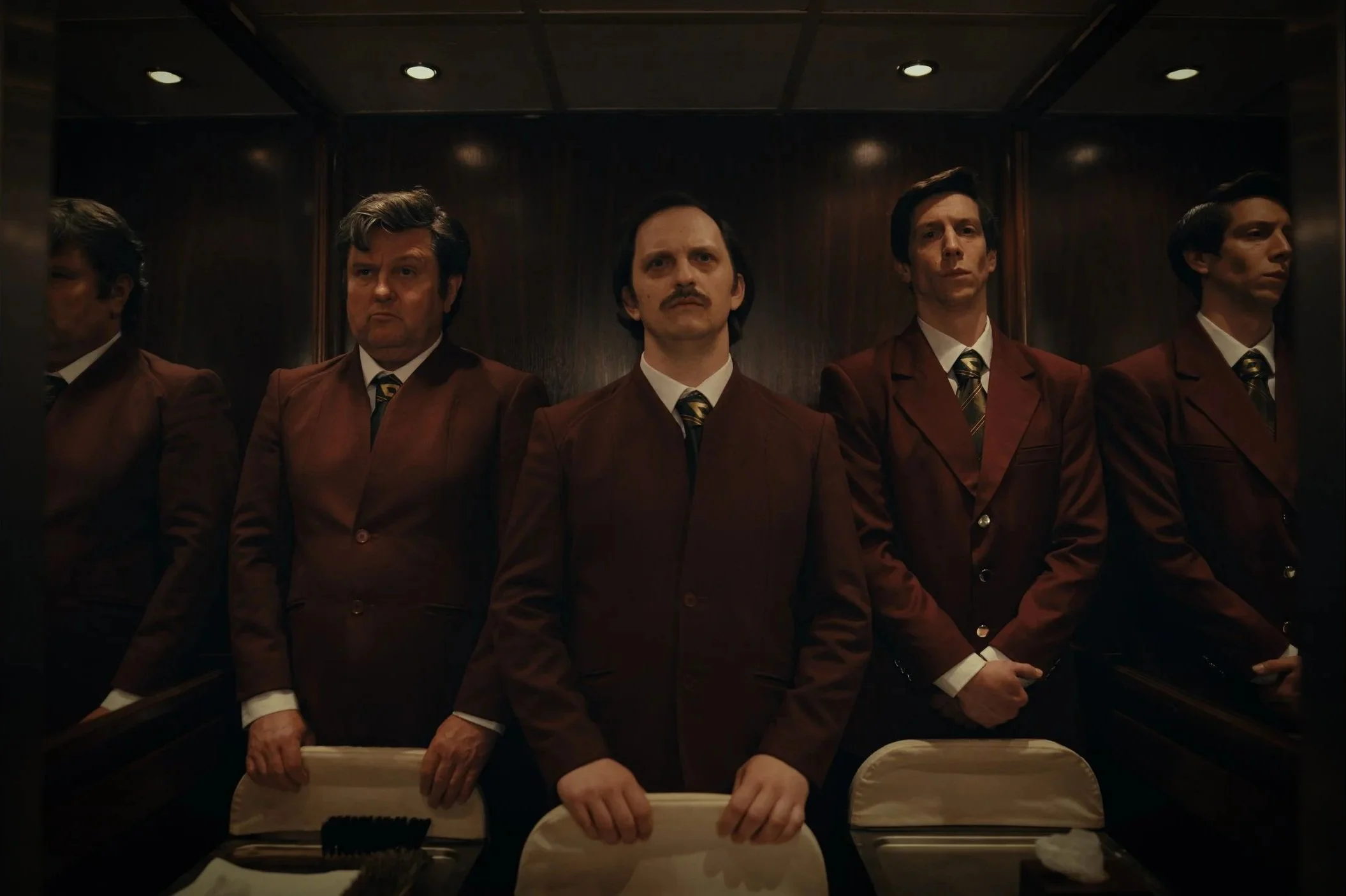 Five men standing in an elevator, dressed in matching brown suits and striped ties, with serious expressions, dark wood paneling in the background.