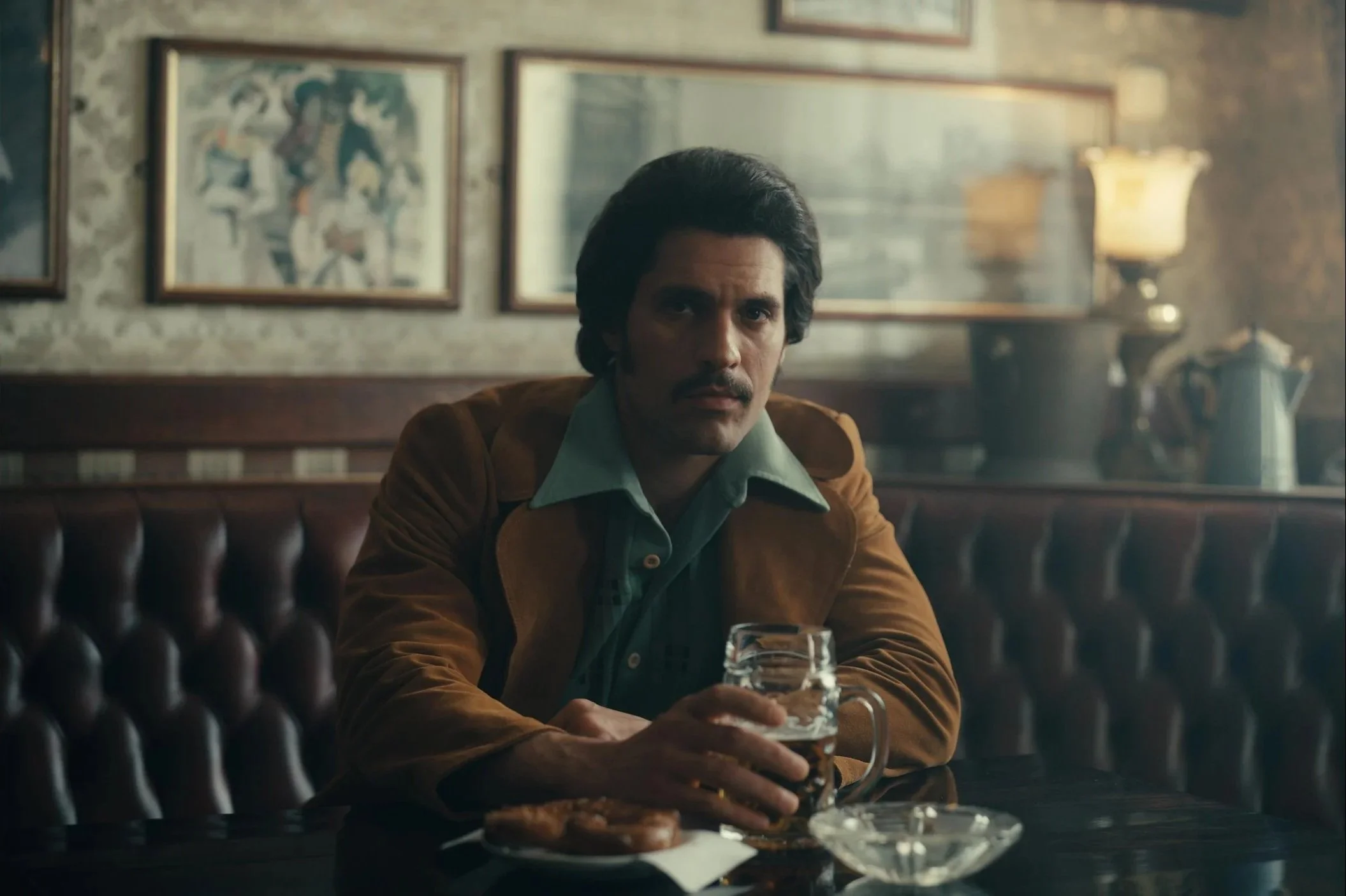 A man sitting alone at a booth in a dimly lit restaurant or bar, holding a glass of dark beverage, with a dessert on the table and framed pictures or artwork on the wall behind him.