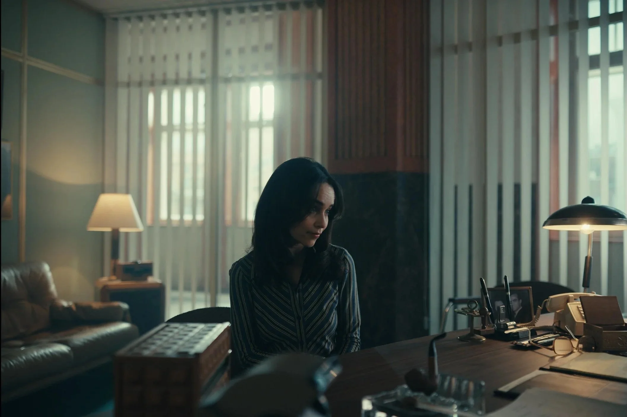 A woman with dark shoulder-length hair sitting at a desk in a dimly lit office, with various office supplies, a telephone, and framed photos on the desk, and large windows with vertical blinds in the background.