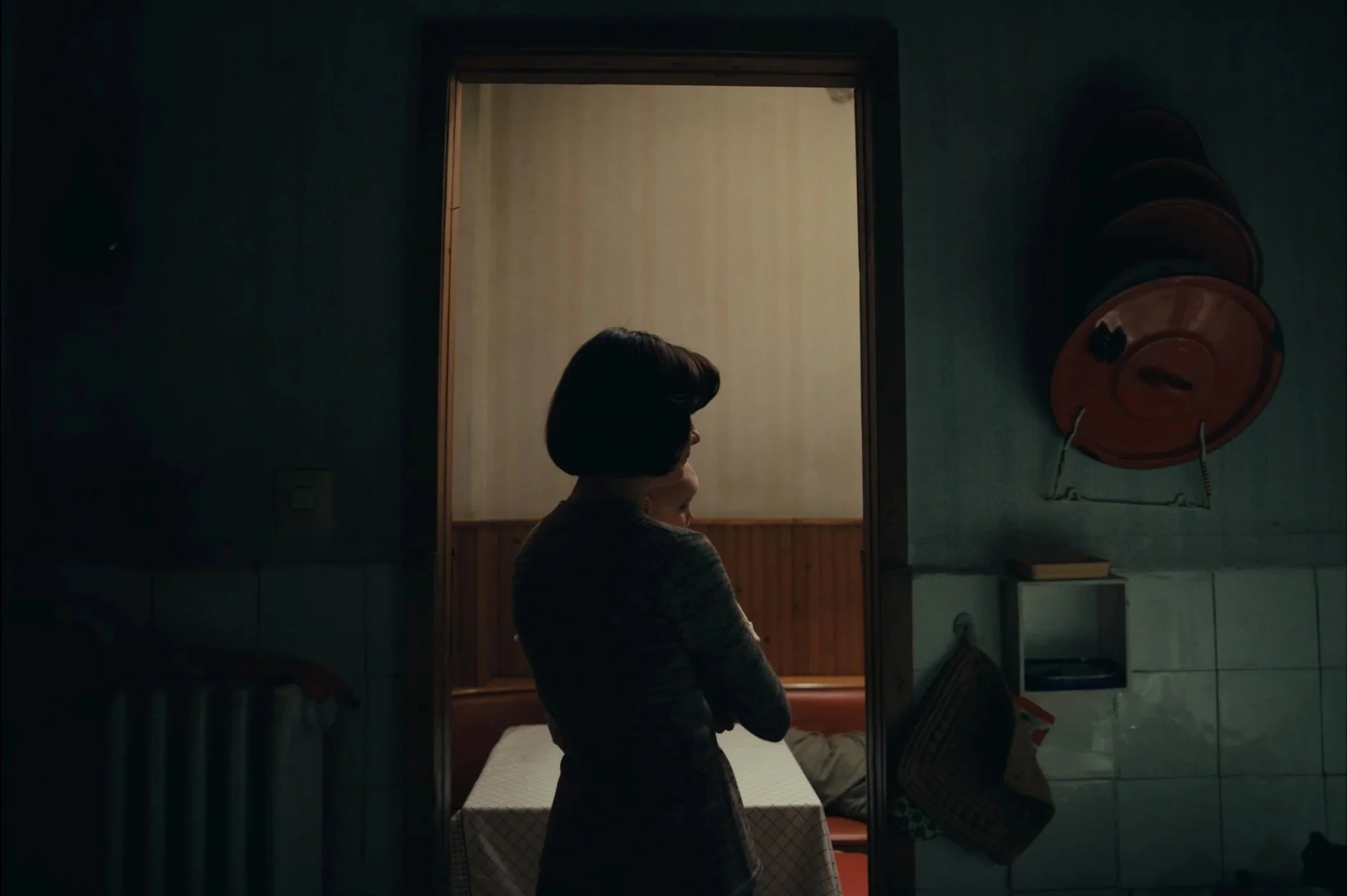 A woman with dark hair and a child standing in a dimly lit room, looking into a brightly lit room through a doorway.