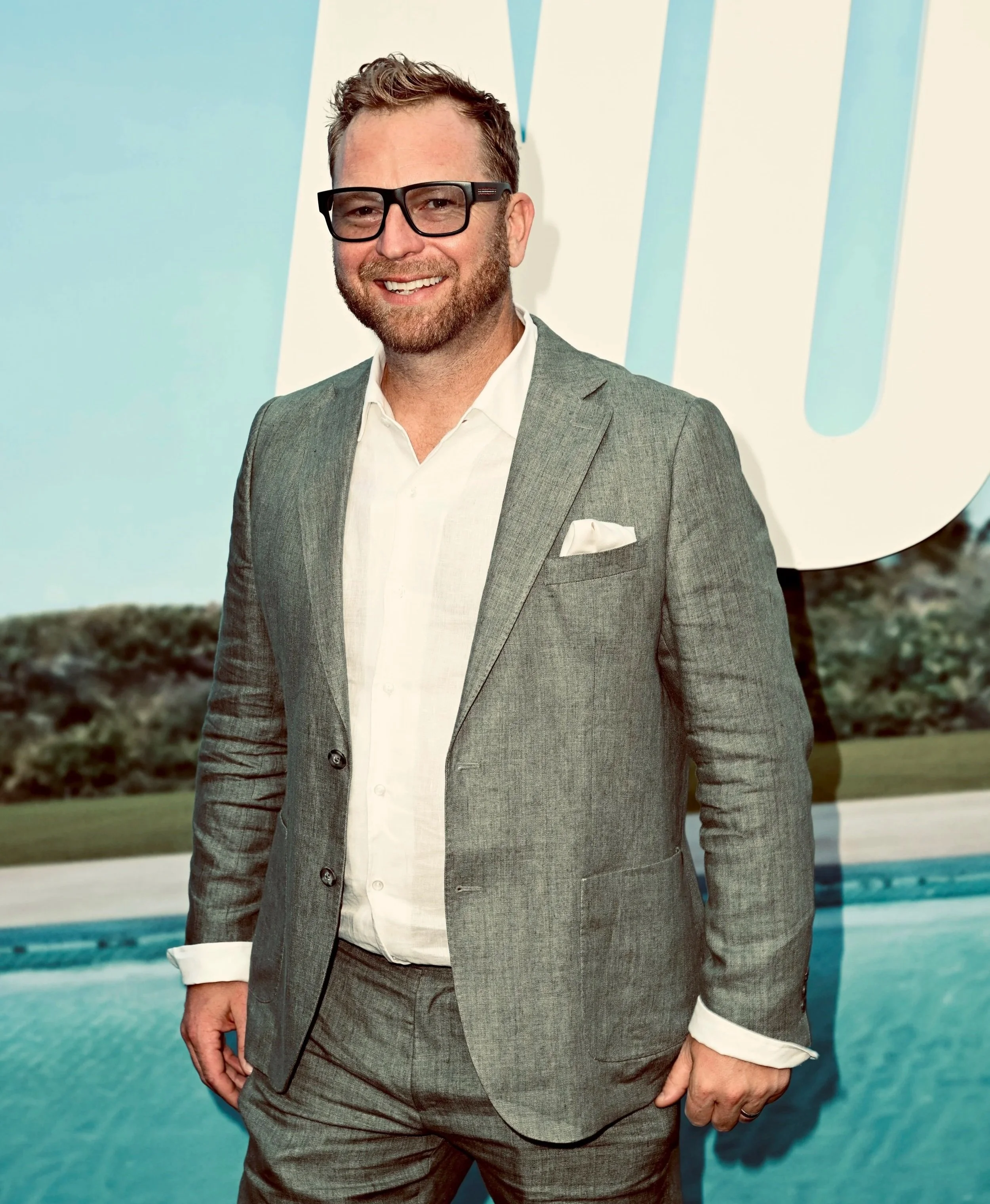 A man in a gray suit with a white shirt, black glasses, and a beard, standing in front of a sea or pool backdrop and smiling.