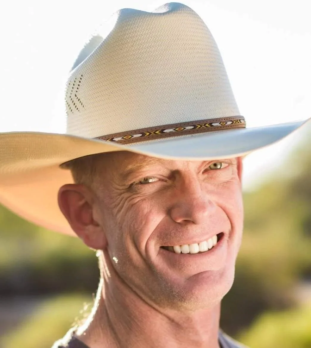 A man smiling outdoors wearing a large white cowboy hat with a decorative band.