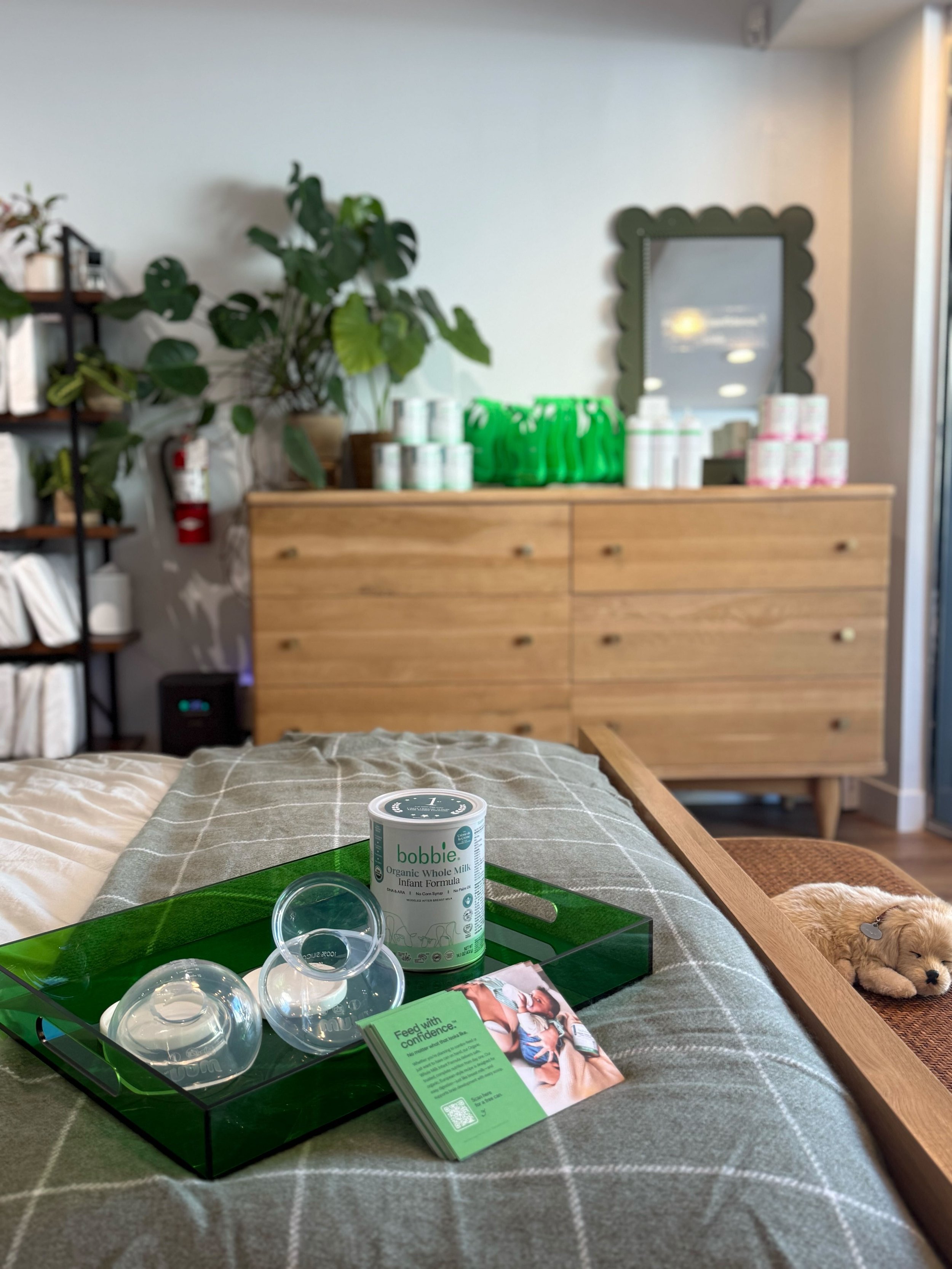A hospital or healthcare setting with a wooden drawer cabinet, various skincare or medical products, a mirror, and a green tray with transparent containers and promotional material in the foreground. There is a large leafy plant and a fire extinguisher on the wall in the background.