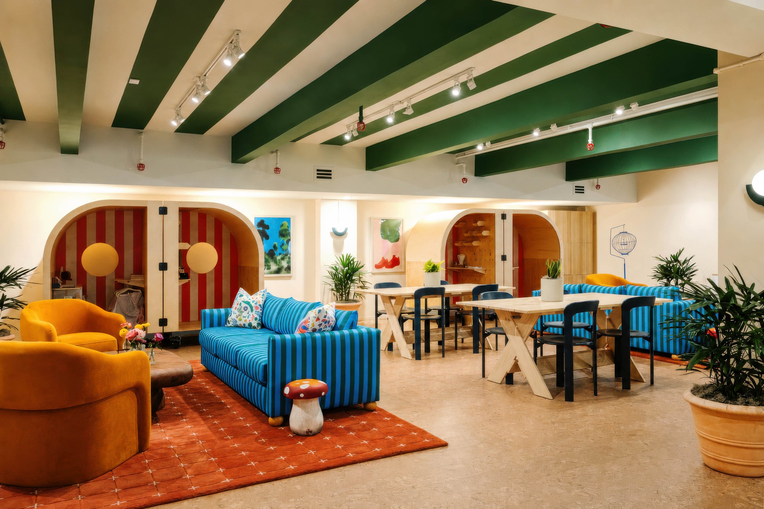 Colorful modern interior of a lounge or common area with striped green and white ceiling beams, potted plants, vibrant blue and yellow sofas, abstract artwork, and dining tables with black chairs.