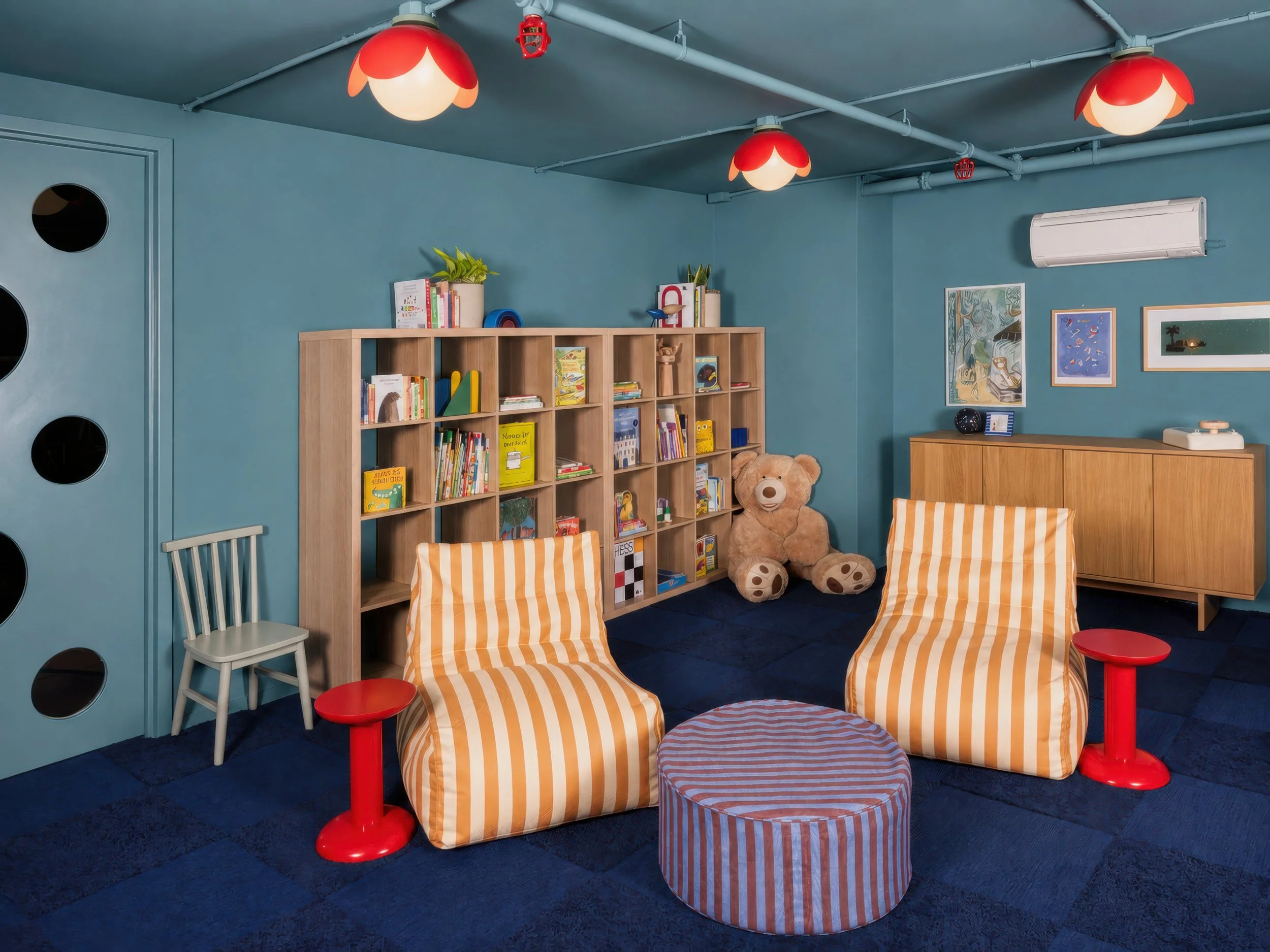 A cozy children's reading corner with a wooden bookshelf filled with colorful books, a large teddy bear sitting beside it, two orange and white striped armchairs, a round ottoman with orange and white stripes, and two small red side tables. The walls are painted blue with framed artwork, and three ceiling lights with red floral-shaped covers hang above. There is an air conditioning unit on the wall.