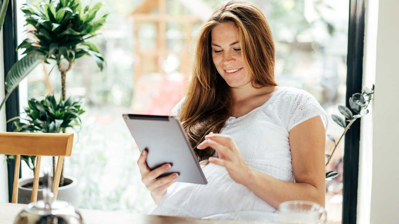 A woman with long red hair, wearing a white shirt, smiling as she uses a tablet in a bright room with large windows and green plants.