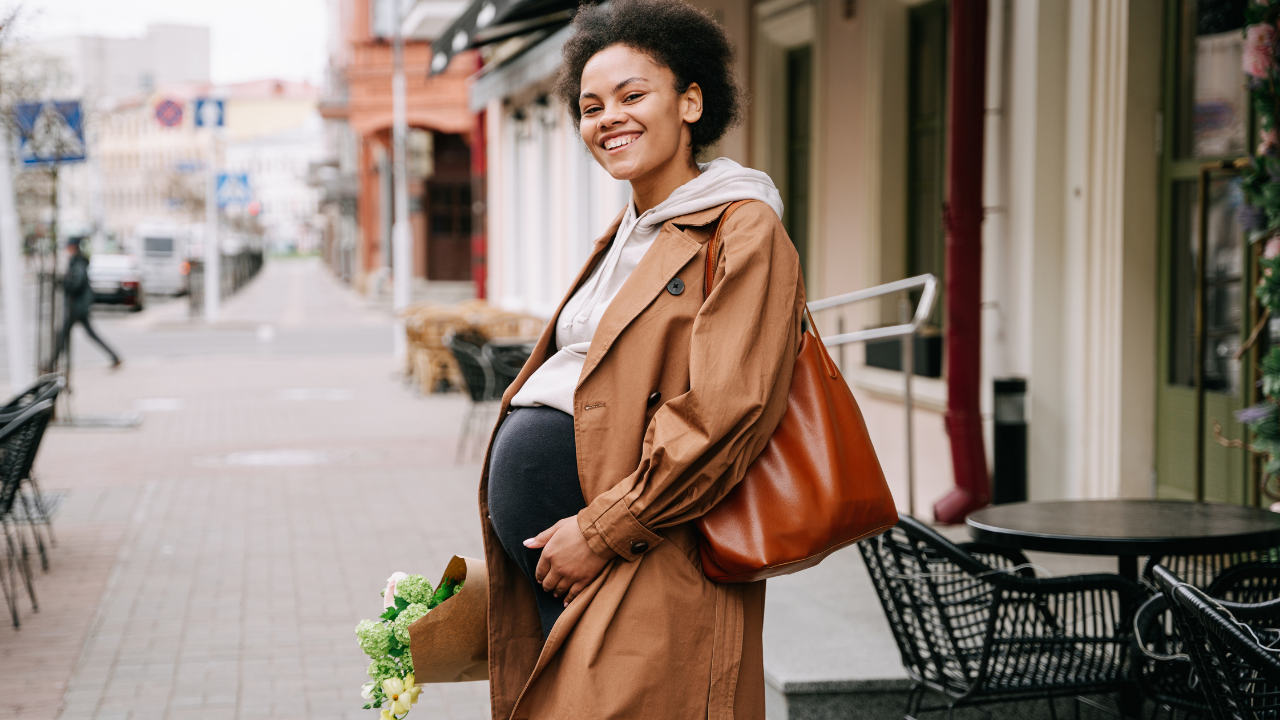 A pregnant woman with curly hair smiling, standing on a city sidewalk, holding a bouquet of flowers, wearing a brown coat and carrying a large brown handbag.