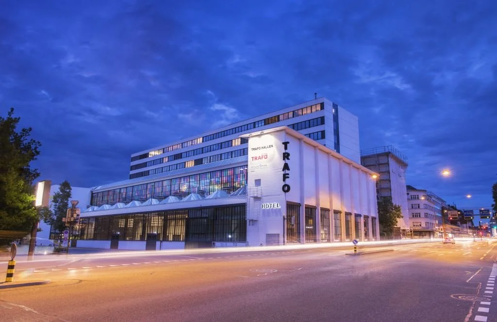 A modern hotel building called Trafo Hallen at dusk, with illuminated signs, glass windows, and traffic light streaks in the street in front.