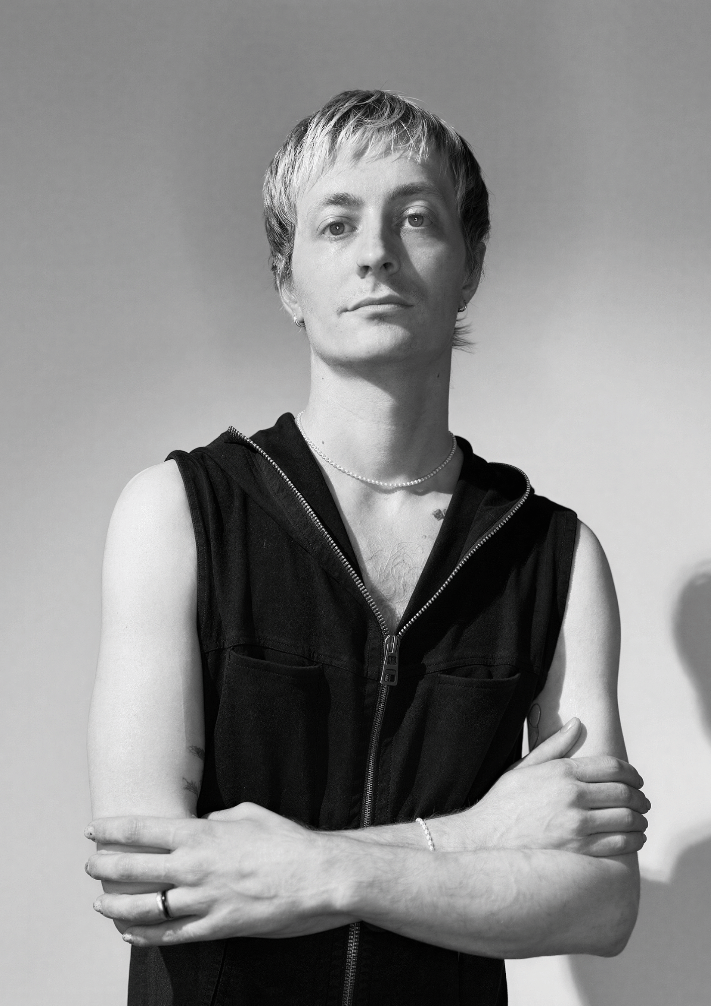 A person with short hair wearing a sleeveless black zip-up vest, standing with arms crossed against a plain background.