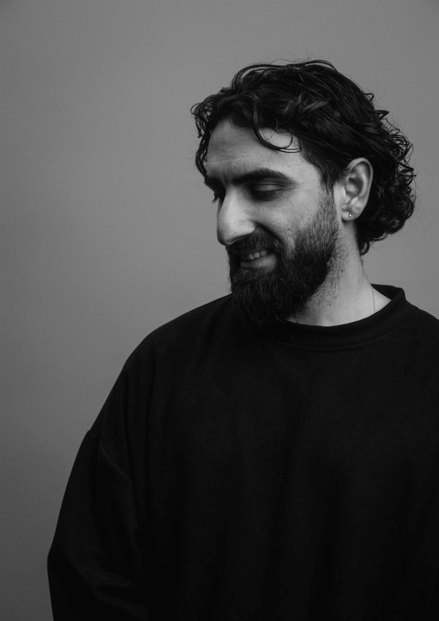 A black and white portrait of a man with dark curly hair and a beard, wearing a dark shirt, looking down with a slight smile