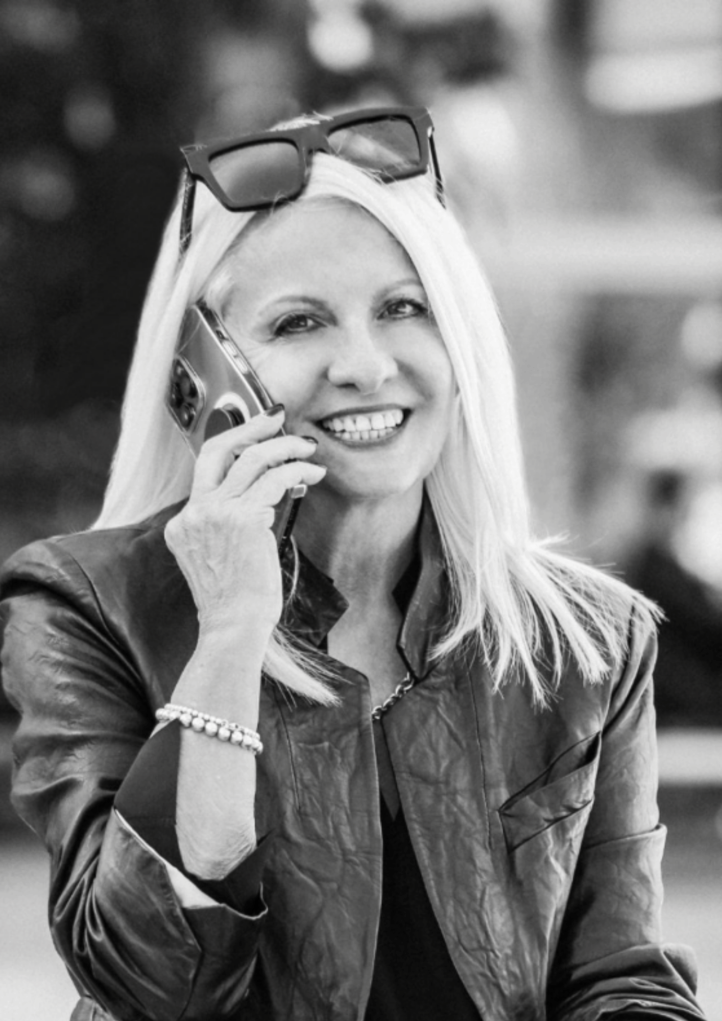 A smiling woman with long blonde hair talking on her cellphone, wearing glasses on her head and a leather jacket.